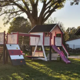 Kolorowy plac zabaw z domkiem, zjeżdżalnią i ścianką wspinaczkową na trawniku w promieniach zachodzącego słońca. Wyraźne cienie drzew dodają głębi.