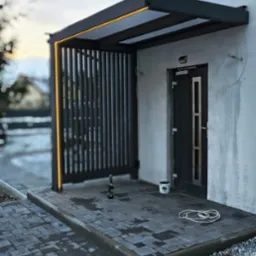 Nowoczesna pergola z pionowymi lamelami i oświetleniem LED przy wejściu do budynku, wykończona kostką brukową. Widoczne narzędzia i wiadro z farbą.