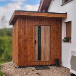 Drewniana, prostokątna konstrukcja z drzwiami, przylegająca do białego budynku z oknami. Na ziemi leżą buty robocze i mata. W tle zieleń.