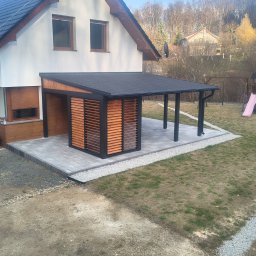 Mariola Starzec - Nowoczesna pergola z drewnianymi żaluzjami, przylegająca do domu, z placem zabaw w tle. Czarna konstrukcja, szare płyty tarasowe i jasne żwirki.
