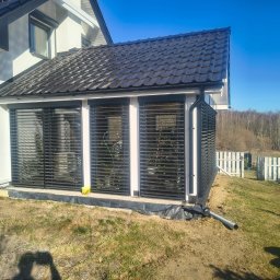 Mariola Starzec - Zadaszona zabudowa balkonu z czarnymi żaluzjami, przylegająca do białego domu z ciemnym dachem. Widoczny fragment ogrodu i białe ogrodzenie w tle.