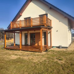 Mariola Starzec - Dwukondygnacyjny taras drewniany przy domu z jasną elewacją. Na parterze zadaszony taras, na piętrze balkon z drewnianą balustradą. Widoczne drewniane słupy i podłoga.