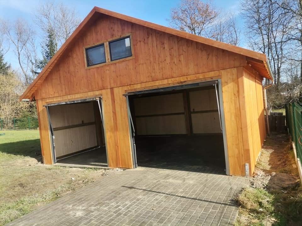 Nowo wybudowany, dwustanowiskowy garaż z drewna, z dwoma oknami na poddaszu, z częściowo otwartymi bramami, na betonowej wylewce.