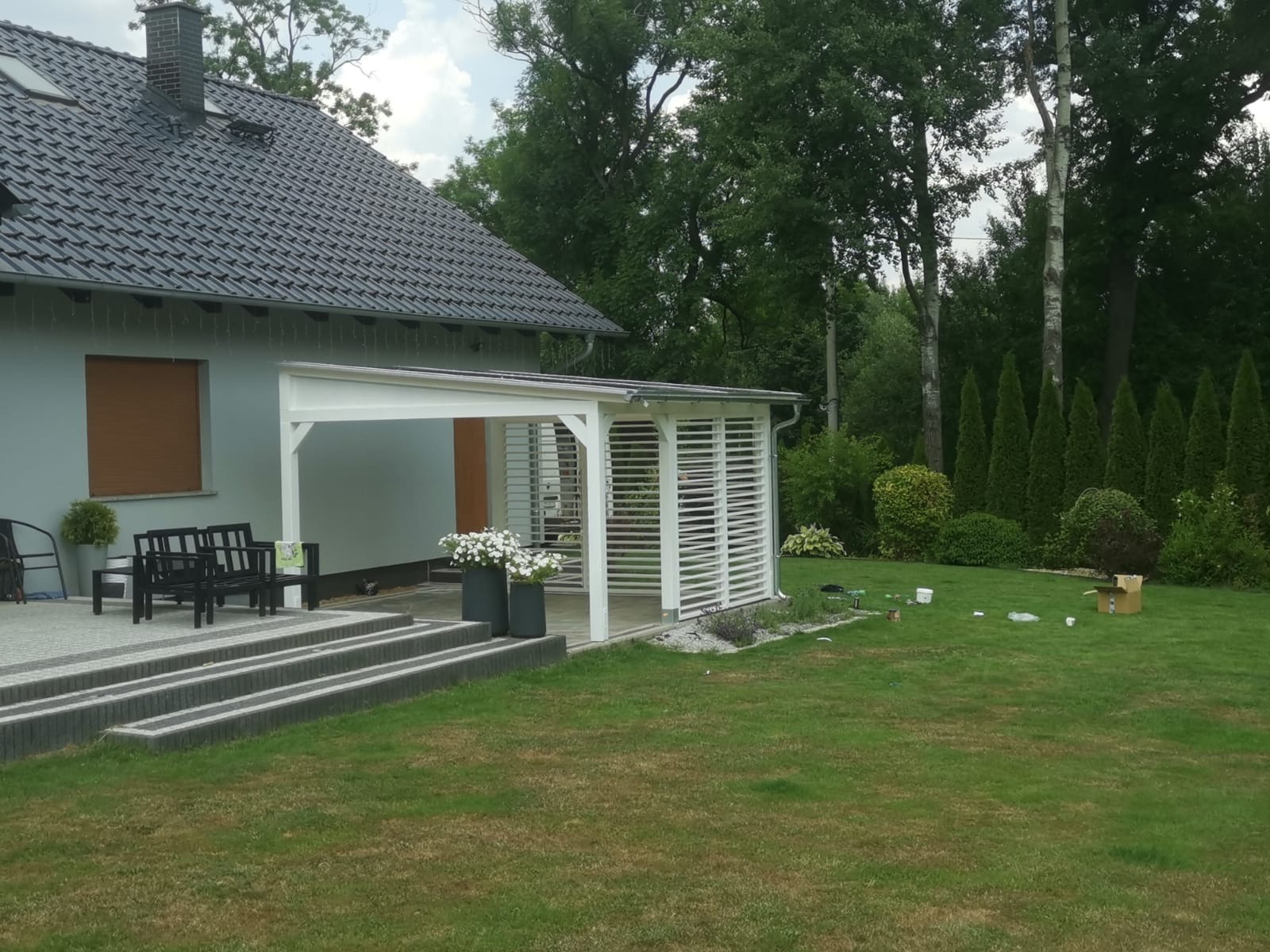 Biała konstrukcja z żaluzjami, prawdopodobnie pawilon ogrodowy, przylegająca do domu z szarą dachówką, z widocznymi schodami, czarnymi ławkami i zielonym trawnikiem z rozrzuconymi śmieciami.