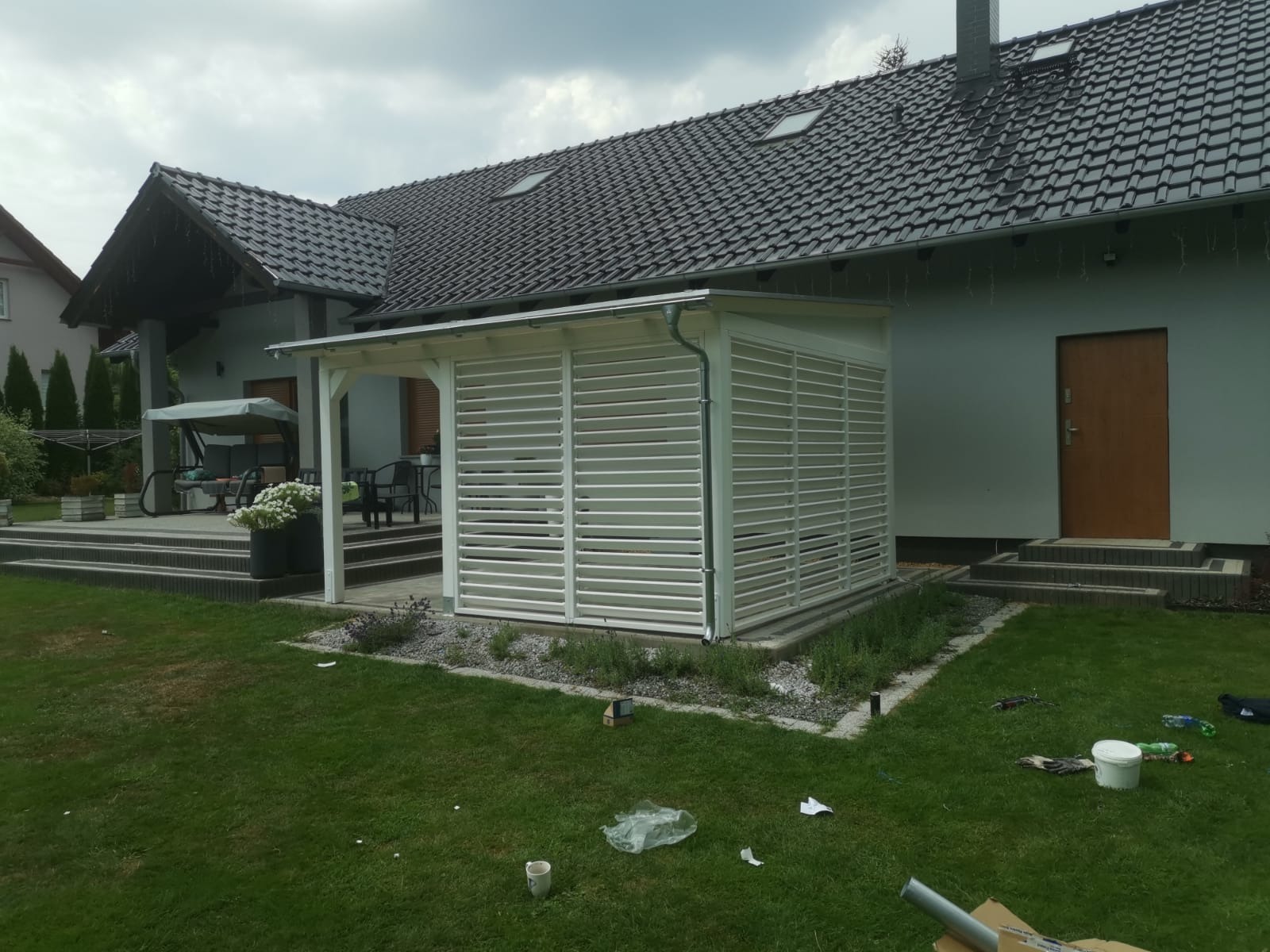 Biała konstrukcja z poziomych listew, przylegająca do domu z ciemną dachówką; widoczne ślady budowy na trawniku.