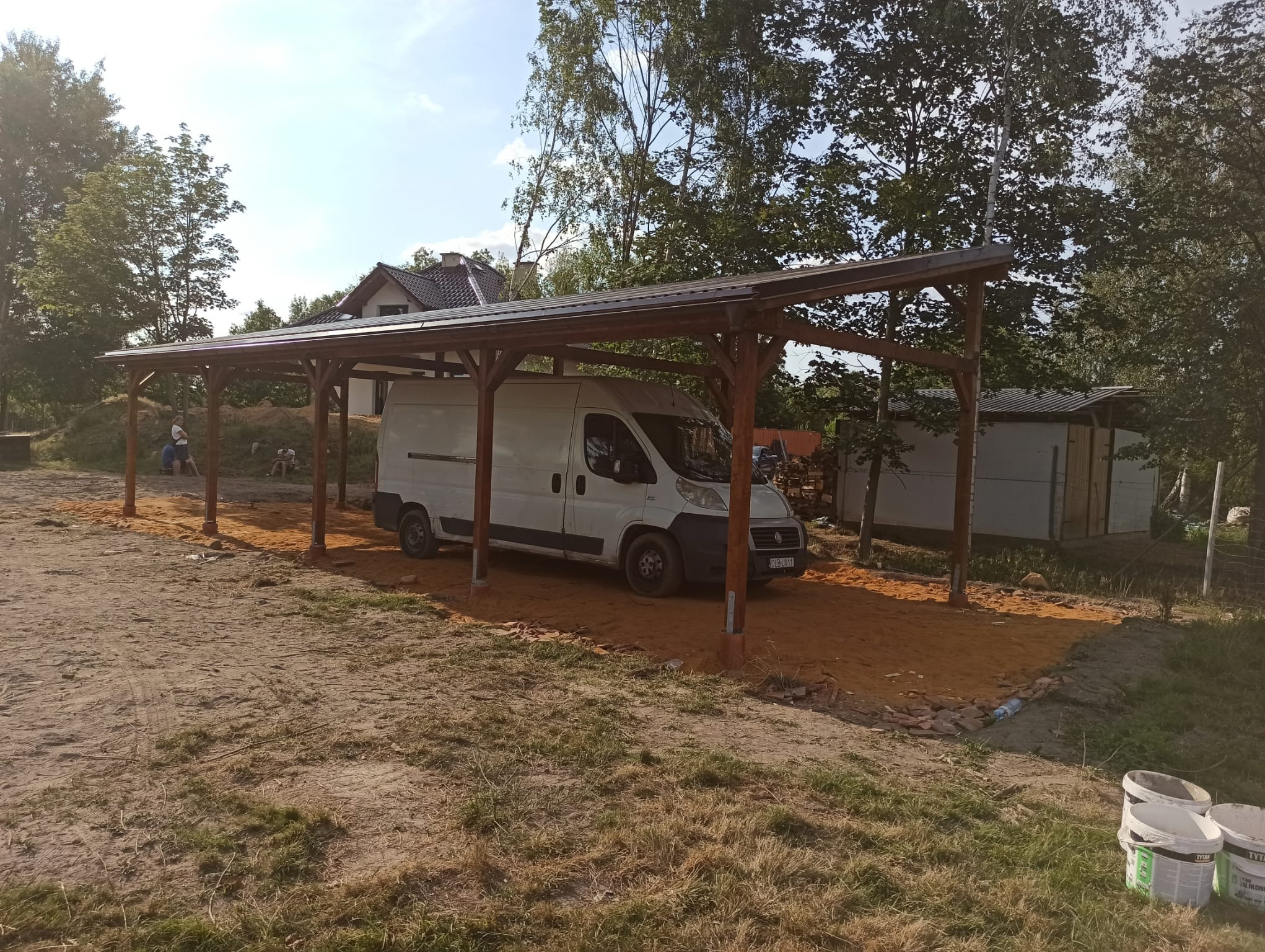 Zadaszona wiata drewniana z brązowym dachem i podłogą z piasku, pod którą stoi biały samochód dostawczy, widoczne drzewa i budynek w tle, na pierwszym planie wiaderka po farbie.