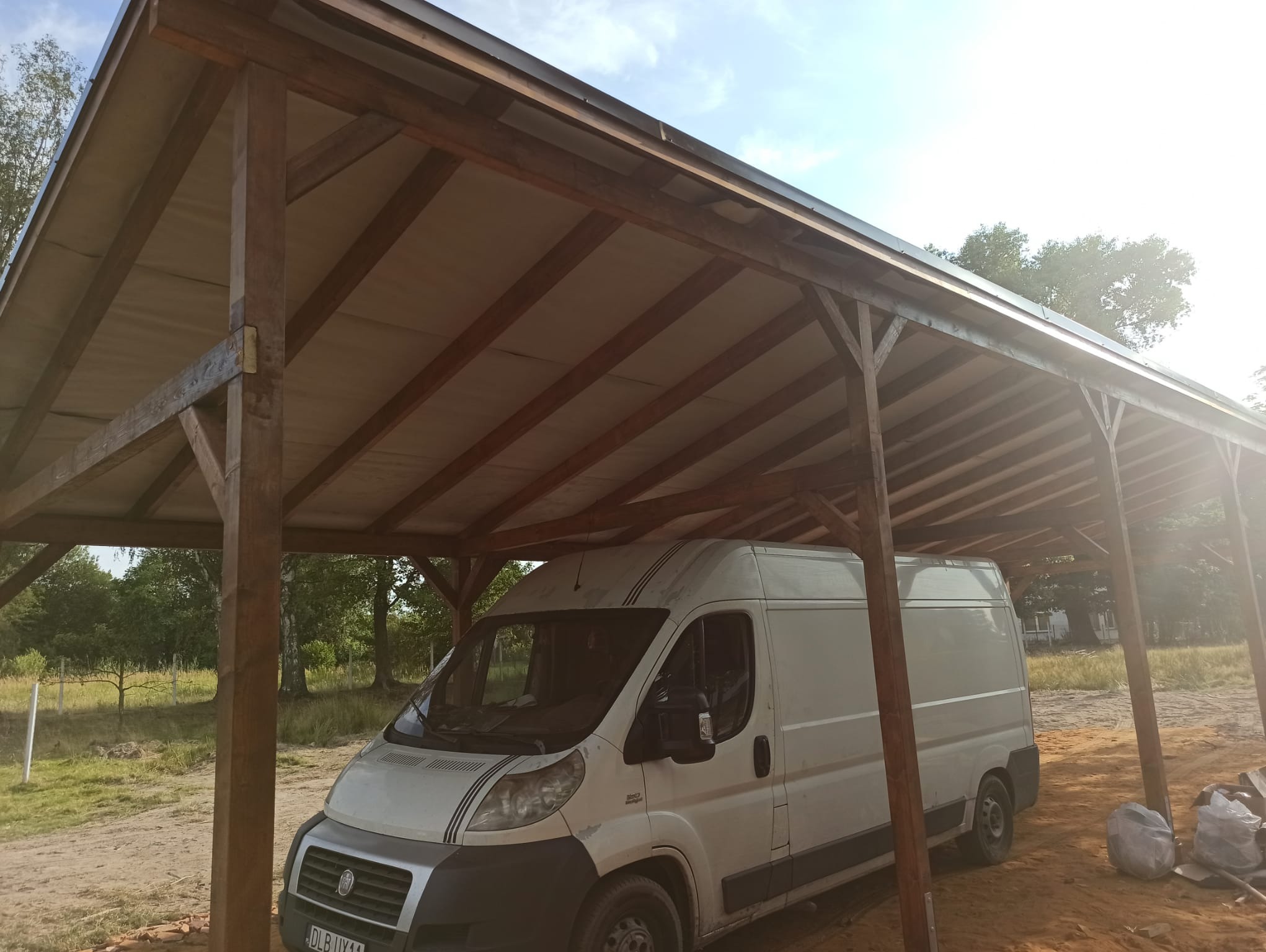 Drewniana konstrukcja z zadaszeniem, pod którą zaparkowany jest biały samochód dostawczy. Widoczne elementy konstrukcyjne i zarys otoczenia.