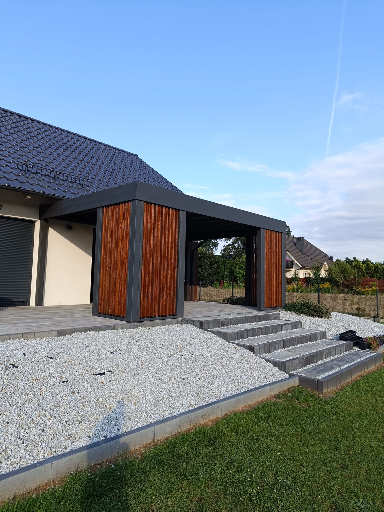 Nowoczesna pergola z drewnianymi panelami i szarą konstrukcją, prowadząca do domu po kamiennych schodach, otoczona białym żwirem i trawnikiem.