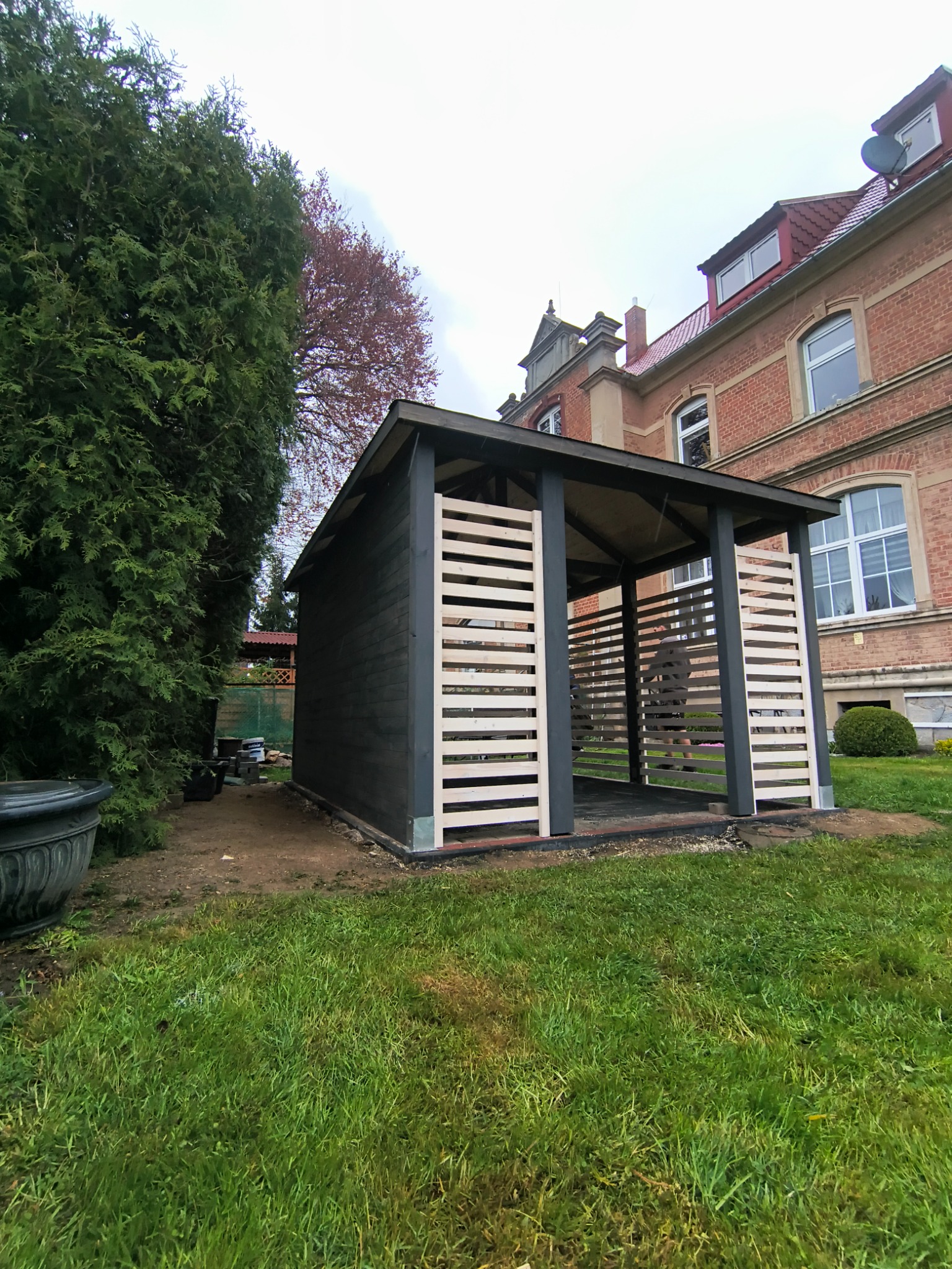 Nowoczesna altana z drewnianymi lamelami w ogrodzie, na tle ceglanego budynku. Konstrukcja pomalowana na ciemny kolor, z jasnymi elementami wykończeniowymi.