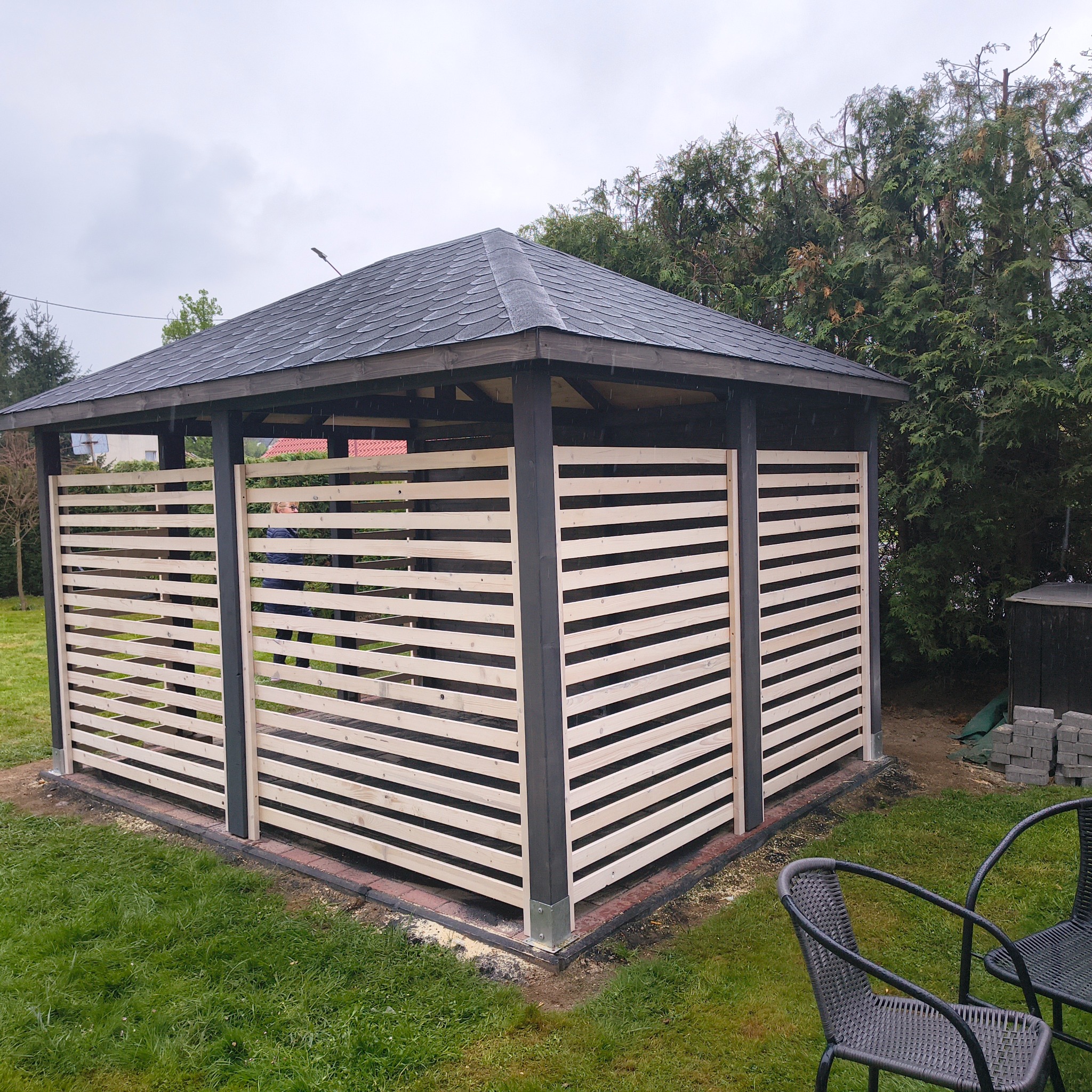 Drewniana altana z szarym dachem i jasnymi, poziomymi deskami na ścianach, otoczona zielonym trawnikiem. W tle drzewa i częściowo widoczne krzesła ogrodowe.
