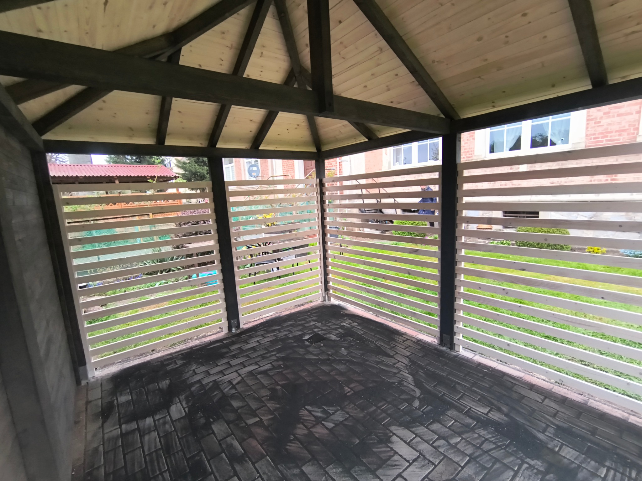 Drewniana altana z ażurowymi ścianami w ogrodzie. Podłoga wyłożona kostką brukową. Widok z wnętrza altany na zieleń ogrodu i budynek z czerwonej cegły.