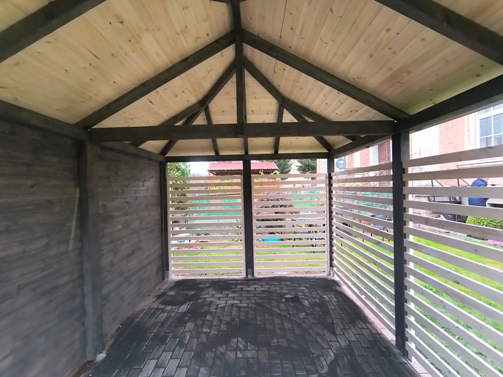 Wnętrze altany z drewnianą konstrukcją, pomalowaną na szaro. Podłoga wyłożona ciemną kostką. Przez ażurowe ściany widać zieleń ogrodu.