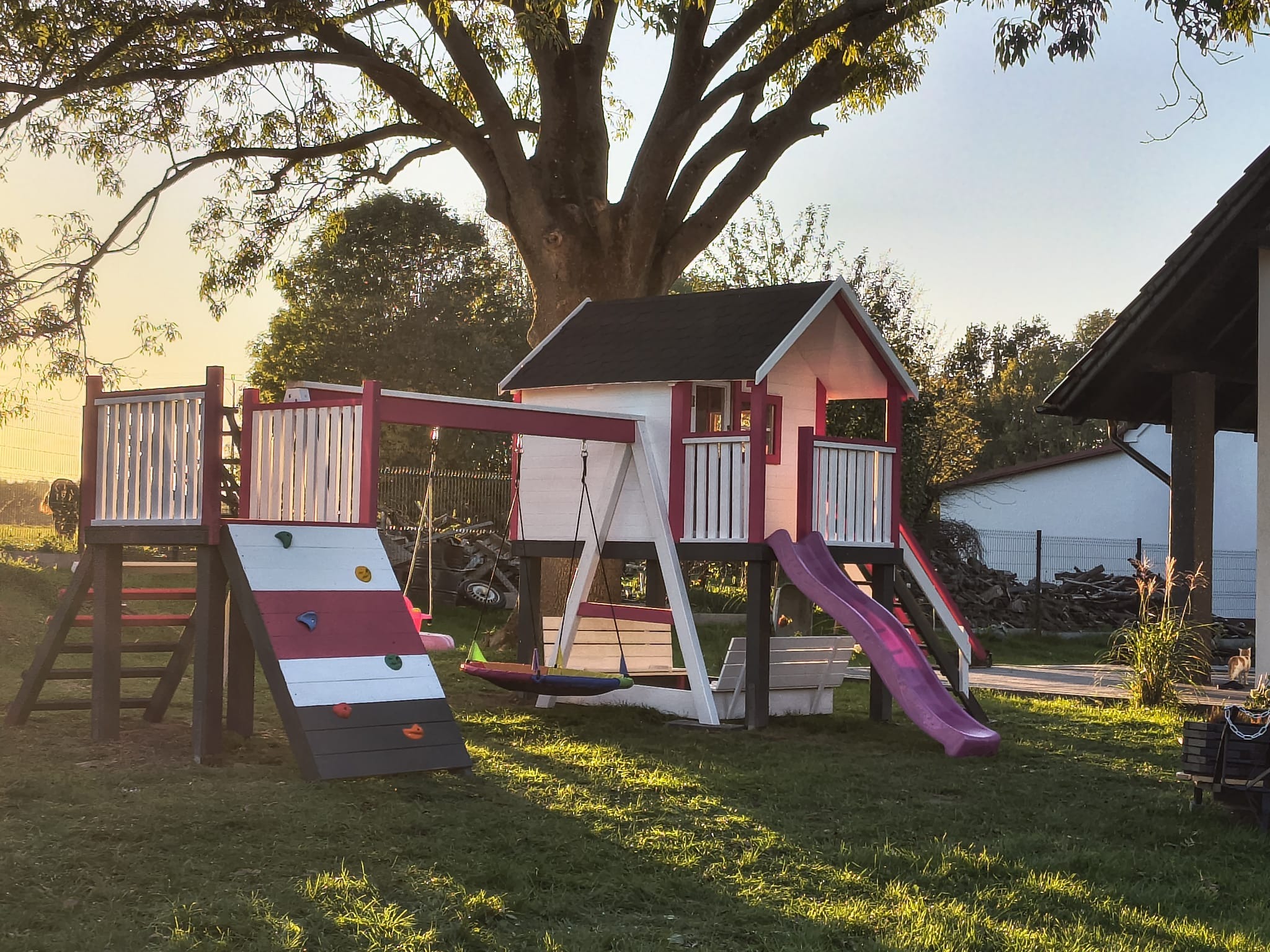 Kolorowy plac zabaw z domkiem, zjeżdżalnią i ścianką wspinaczkową na trawniku w promieniach zachodzącego słońca. Wyraźne cienie drzew dodają głębi.