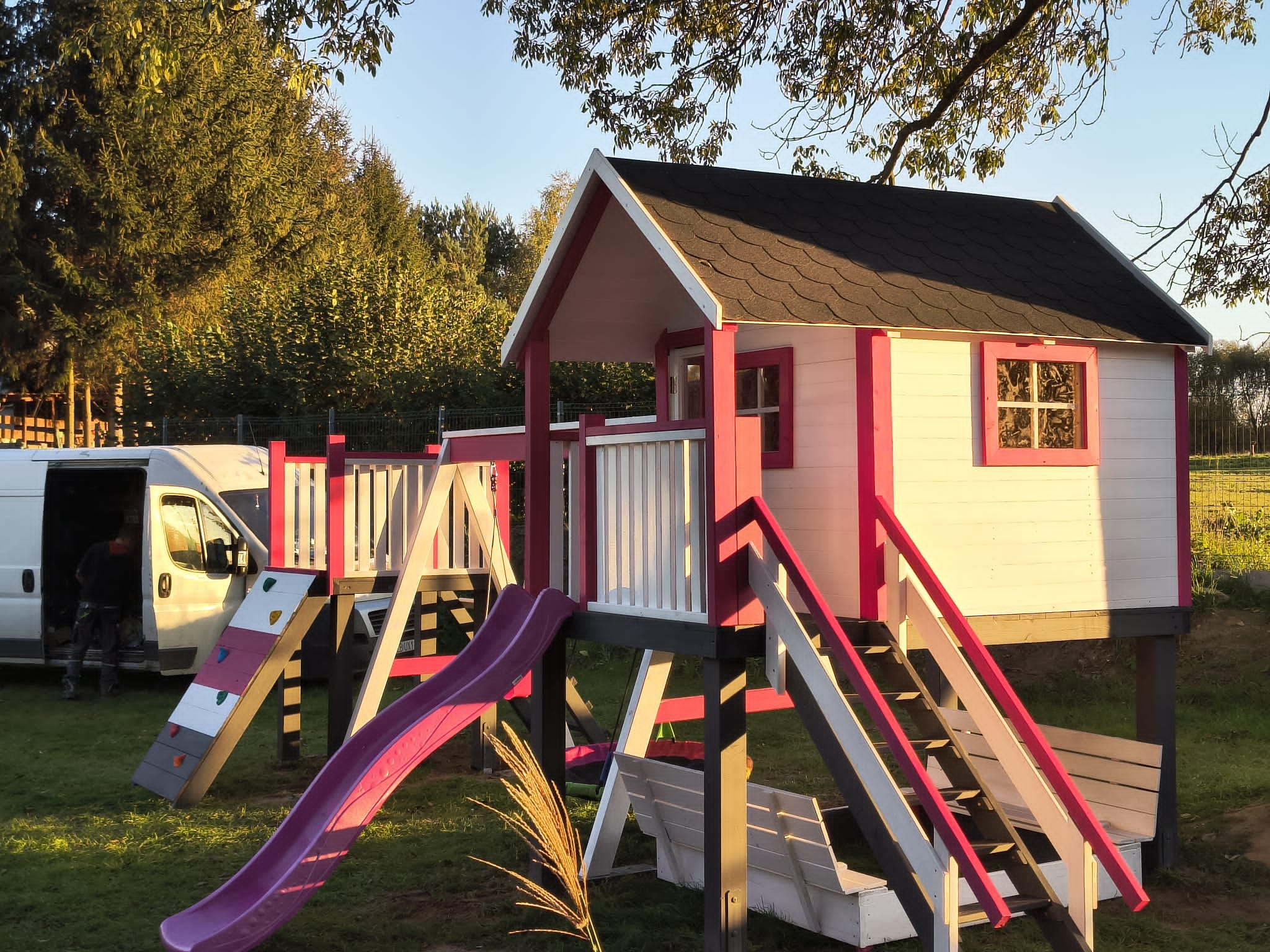 Kolorowy plac zabaw z domkiem na drzewie, zjeżdżalnią i ścianką wspinaczkową, ustawiony na trawniku. W tle częściowo widoczny bus dostawczy.