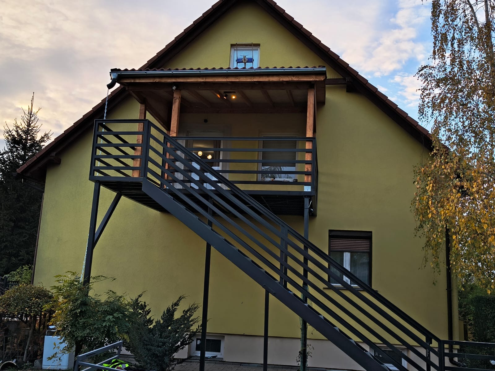 Zewnętrzne schody metalowe prowadzące na balkon z drewnianą konstrukcją w Olszynie. Widok na elewację domu z oknami i fragmentem ogrodu.