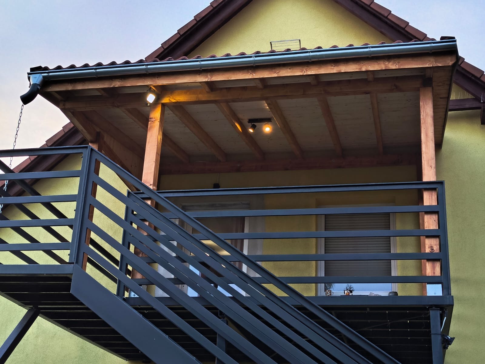 Zadaszony balkon z metalowymi schodami i balustradą w kolorze grafitowym. Widoczne drewniane elementy konstrukcyjne z oświetleniem.