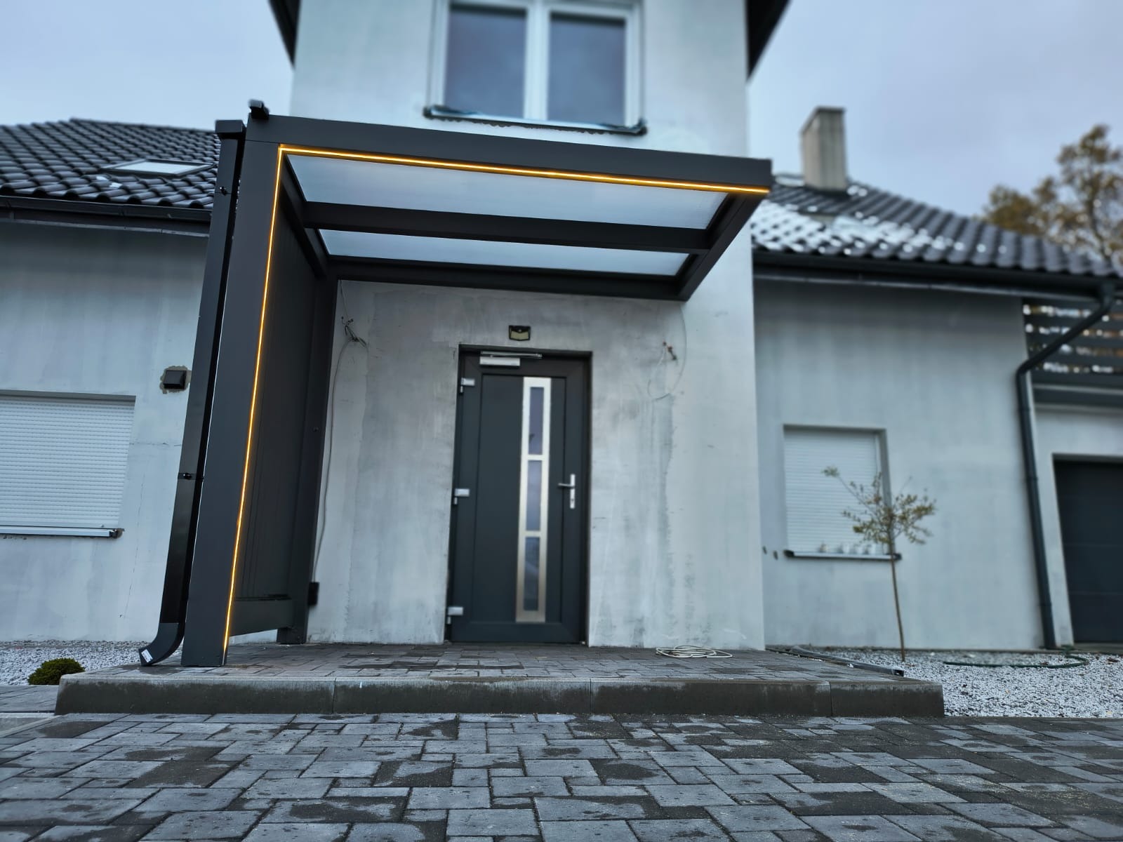 Nowoczesna, minimalistyczna pergola przy wejściu do domu, oświetlona ledowym światłem, z szarą kostką brukową i elewacją w tle.
