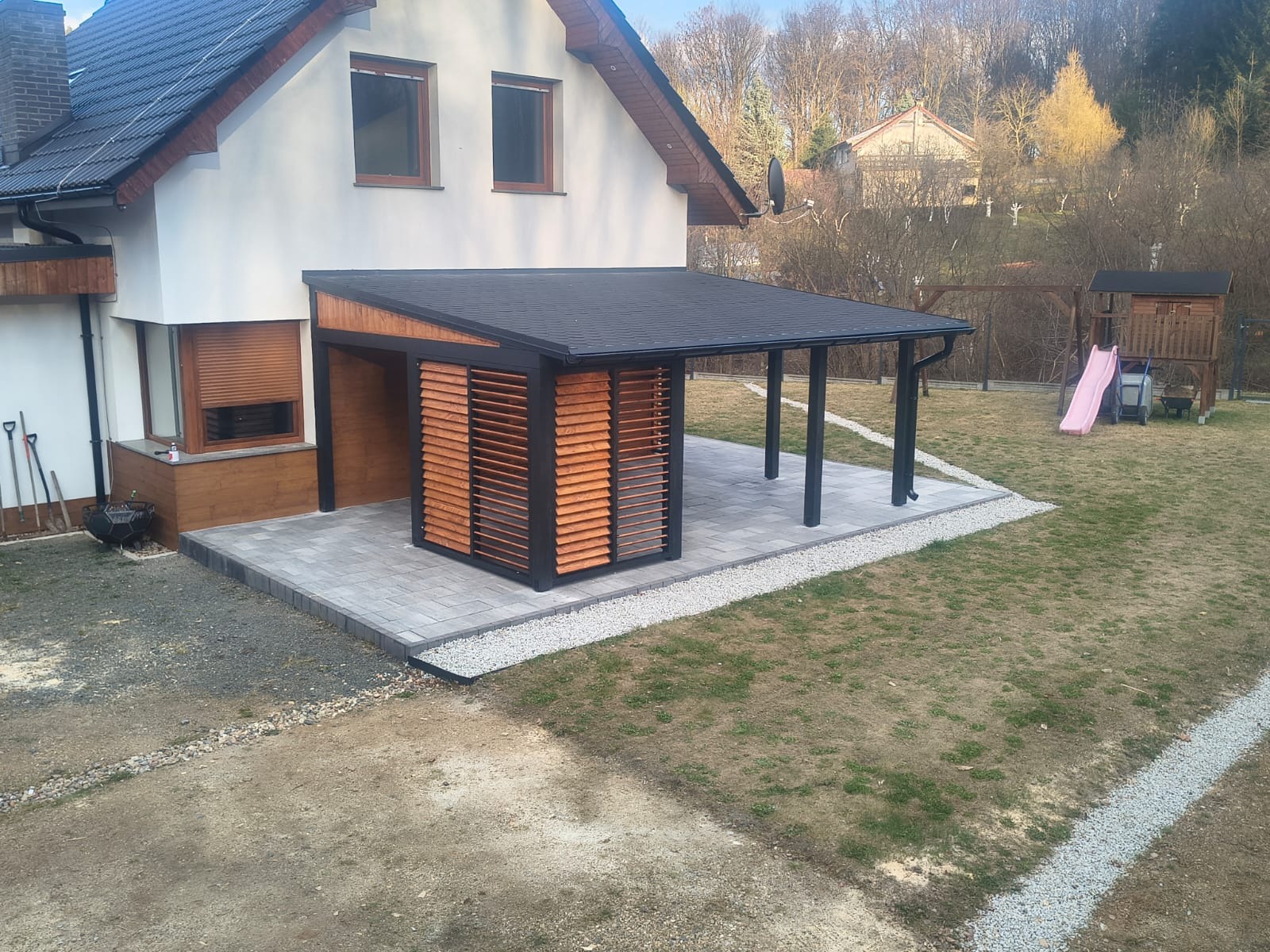 Nowoczesna pergola z drewnianymi żaluzjami, przylegająca do domu, z placem zabaw w tle. Czarna konstrukcja, szare płyty tarasowe i jasne żwirki.