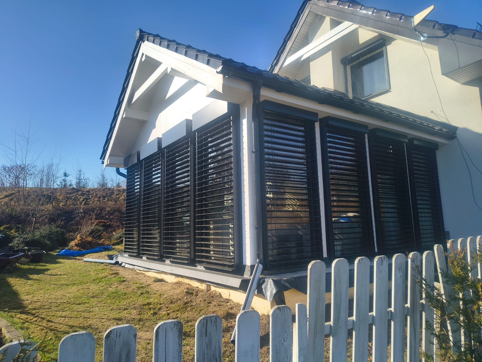 Zabudowa tarasu z żaluzjami fasadowymi w kolorze antracytowym, widok z zewnątrz przez białe, drewniane ogrodzenie. Jasne tło budynku i błękitne niebo.
