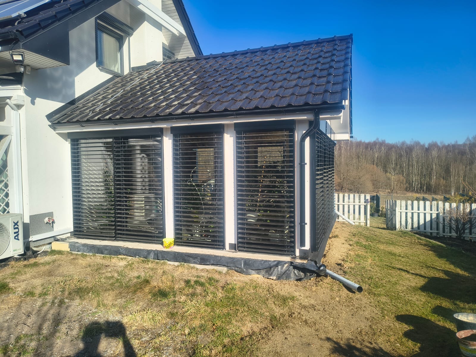 Zadaszona zabudowa balkonu z czarnymi żaluzjami, przylegająca do białego domu z ciemnym dachem. Widoczny fragment ogrodu i białe ogrodzenie w tle.