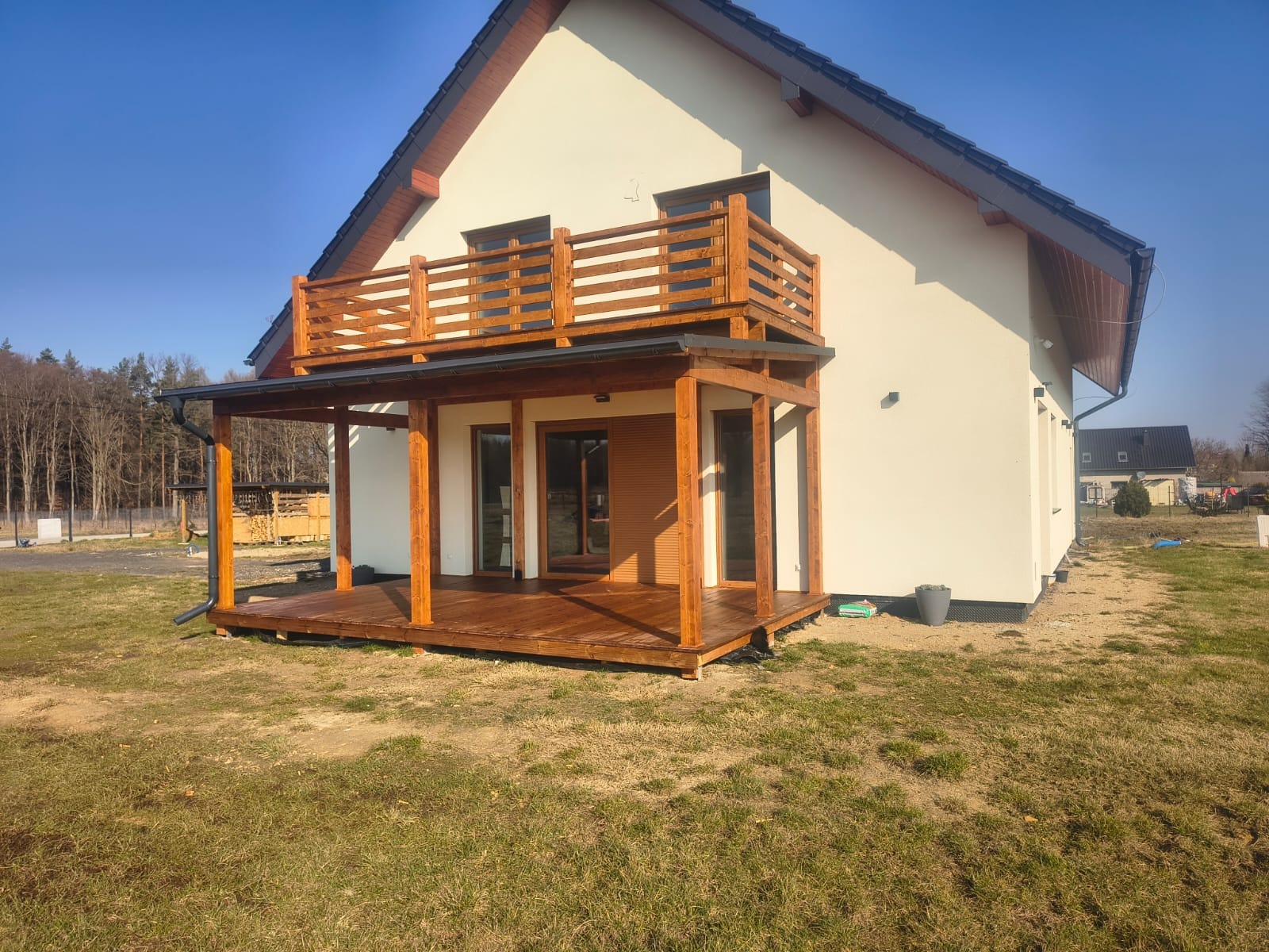 Dwukondygnacyjny taras drewniany przy domu z jasną elewacją. Na parterze zadaszony taras, na piętrze balkon z drewnianą balustradą. Widoczne drewniane słupy i podłoga.