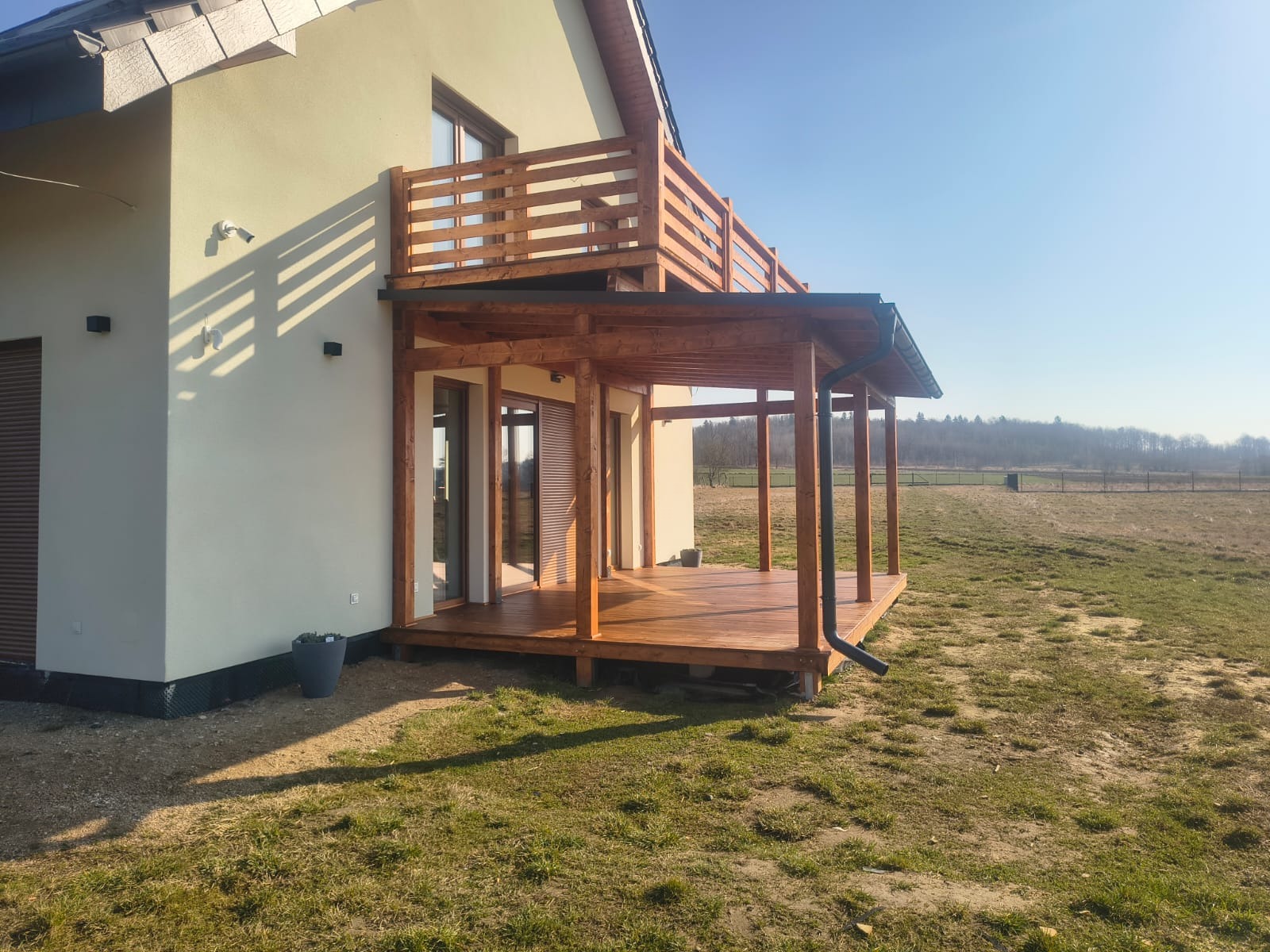 Drewniany taras z zadaszeniem przy domu jednorodzinnym, z widocznym balkonem na piętrze. Konstrukcja z belek, brązowa podłoga tarasu, rynna odprowadzająca wodę.