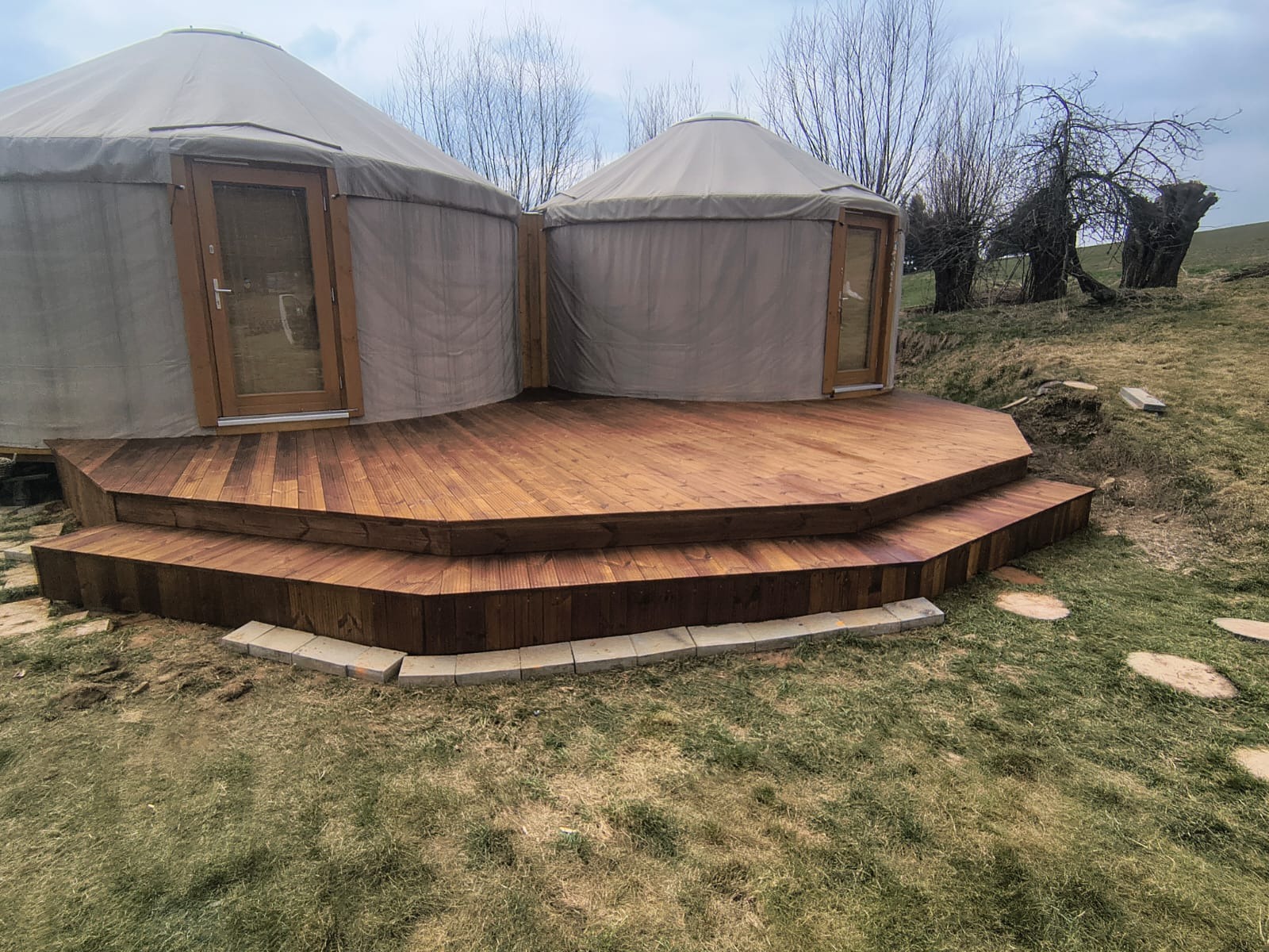 Drewniany taras z dwoma poziomami schodów prowadzący do dwóch namiotów jurta. Taras wykończony kamienną obwódką. Widok na wzgórze i drzewa w tle.