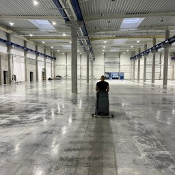 BETA SYSTEM POSADZKI PRZEMYSŁOWE - Pracownik obsługuje maszynę czyszczącą posadzkę w przestronnej hali przemysłowej z betonowymi filarami i oświetleniem sufitowym, widoczne ślady czyszczenia na szarej powierzchni.