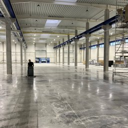 BETA SYSTEM POSADZKI PRZEMYSŁOWE - Duża hala przemysłowa z betonową posadzką w trakcie mycia maszyną czyszczącą, widoczne ślady po czyszczeniu, wysokie sufity z oknami dachowymi i betonowe kolumny.