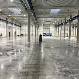 BETA SYSTEM POSADZKI PRZEMYSŁOWE - Pracownik obsługuje maszynę czyszczącą na dużej, szarej powierzchni w hali przemysłowej z betonowymi kolumnami i świetlikami w suficie.