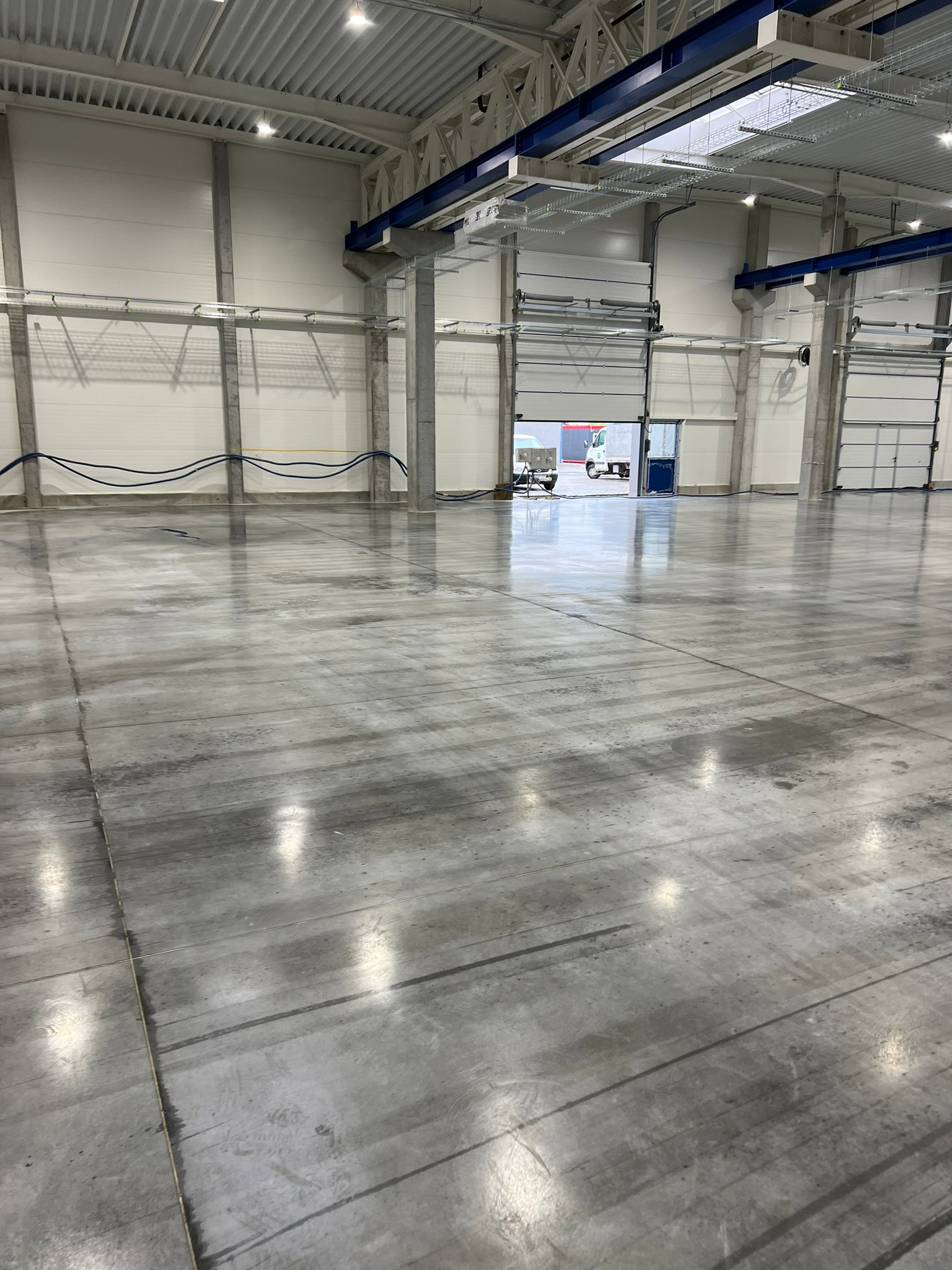 Gładka, szara posadzka w hali przemysłowej z widocznymi bramami wjazdowymi i instalacjami pod sufitem.