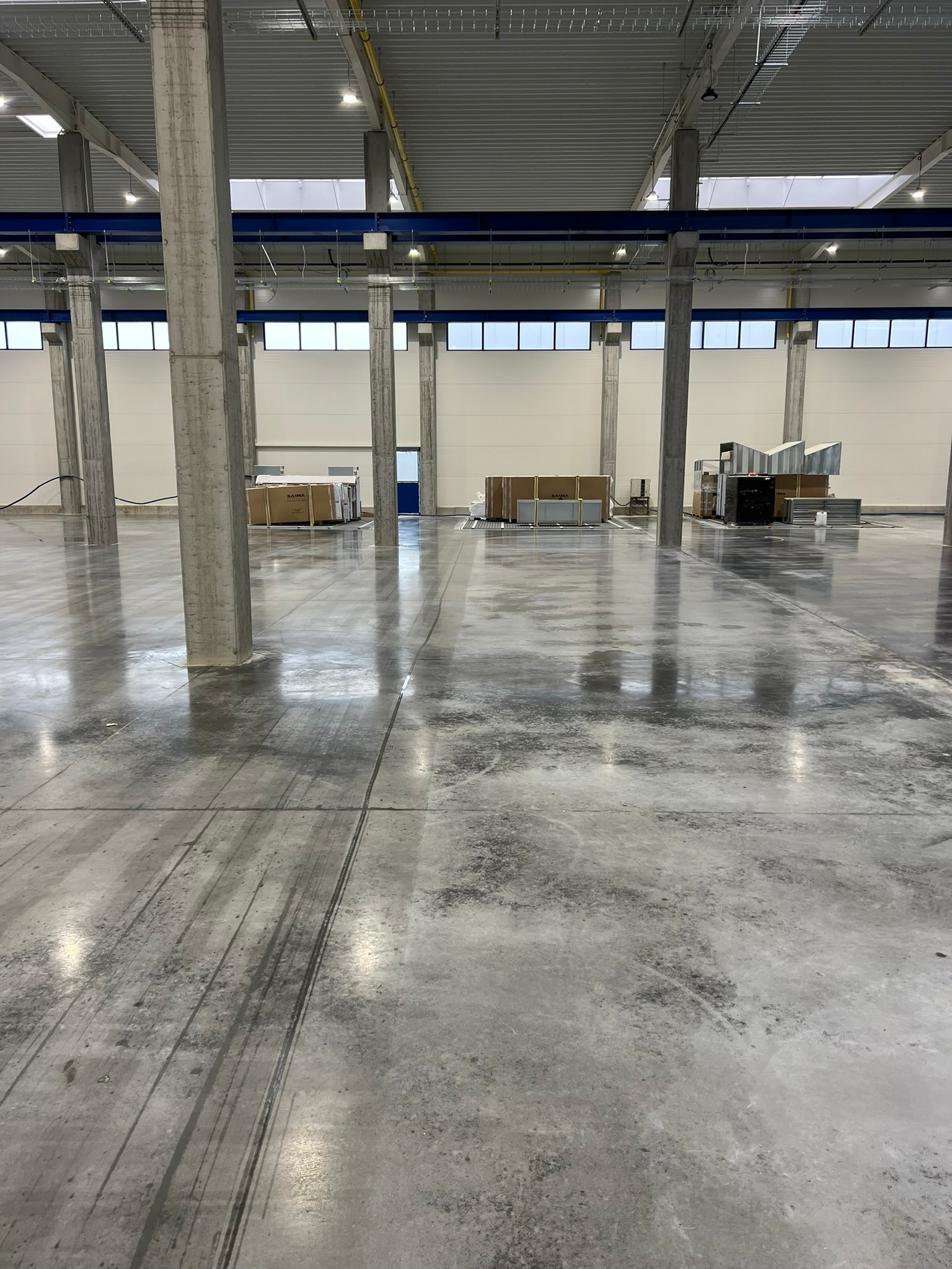 Wnętrze hali przemysłowej z betonową posadzką, widoczne betonowe kolumny, okna w górnej części ścian i ślady opon na podłodze.