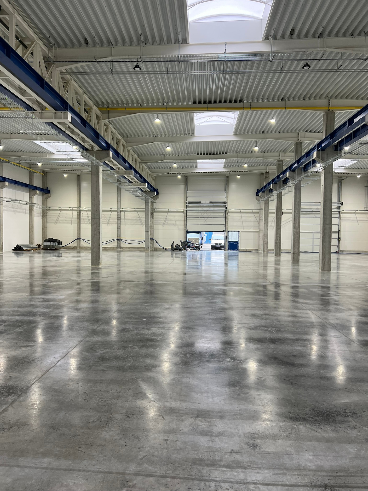 Nowa, gładka, szara posadzka w hali przemysłowej z widocznymi słupami nośnymi i oświetleniem sufitowym.