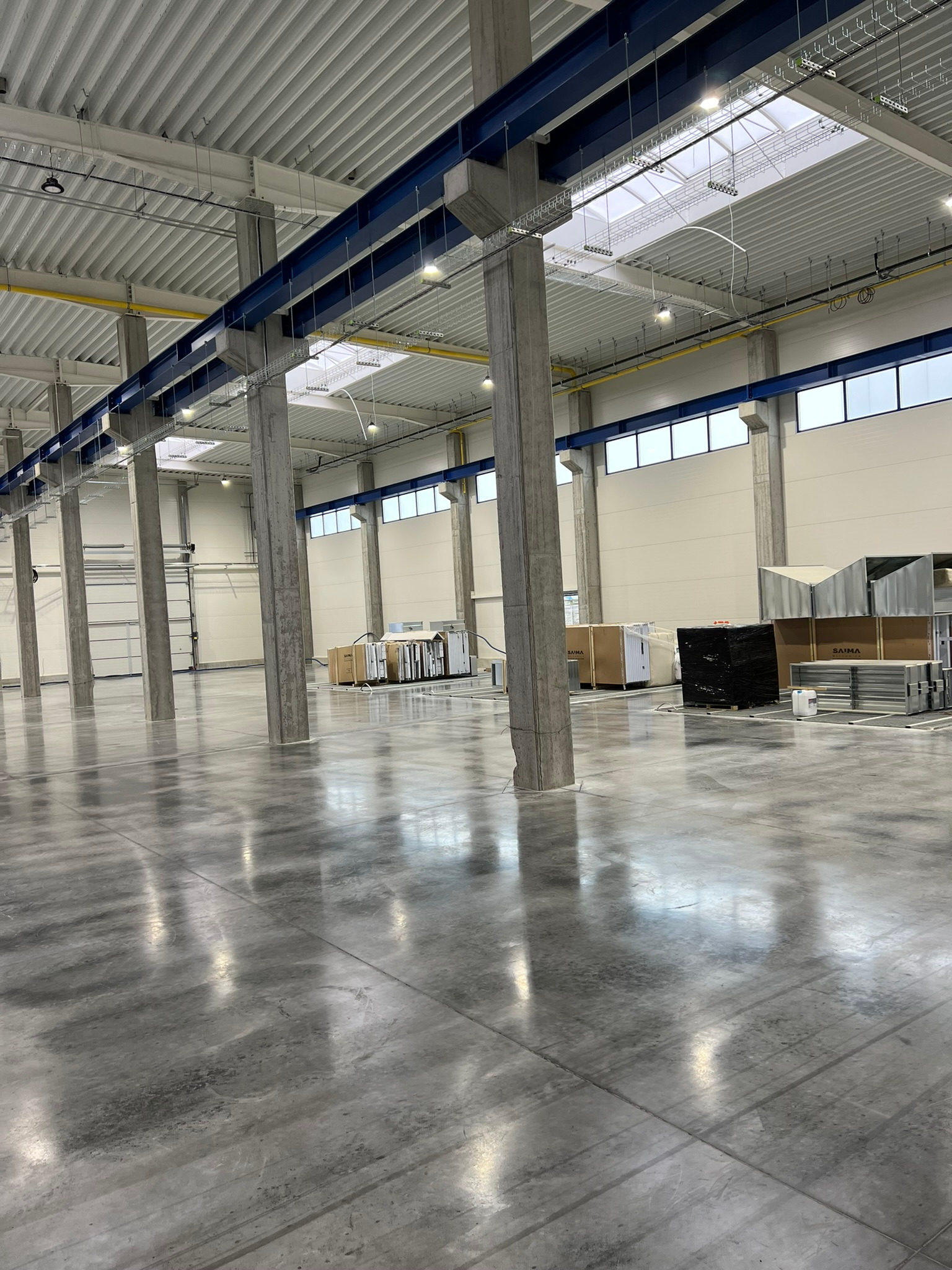 Wnętrze hali przemysłowej z gładką, szarą posadzką, betonowymi kolumnami i niebieskimi belkami pod sufitem. Światło wpada przez okna i świetliki dachowe, odbijając się w posadzce. Widoczne palety...