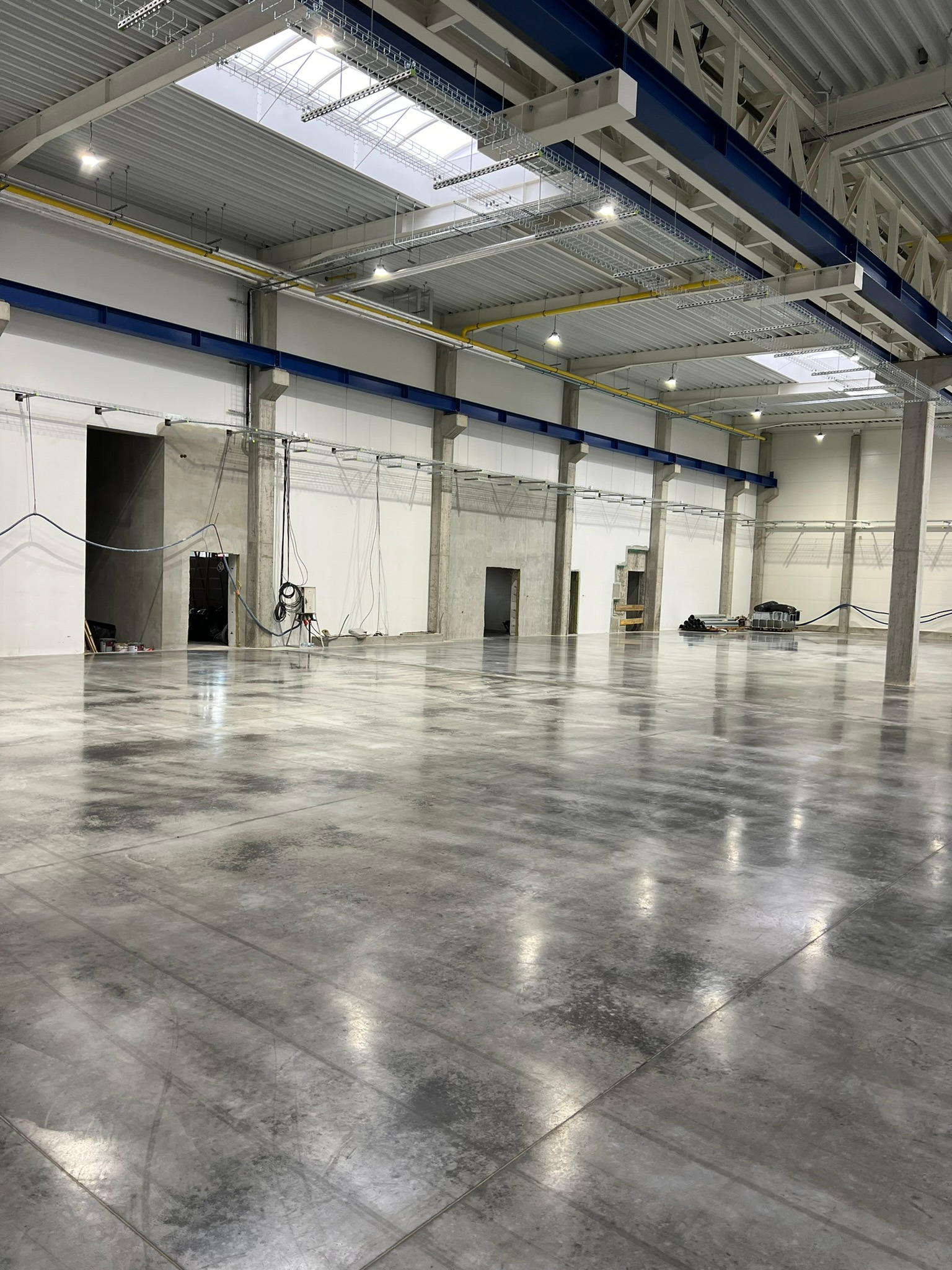 Wnętrze hali przemysłowej z gładką, szarą posadzką. Widoczne betonowe słupy, metalowa konstrukcja dachu z oknami doświetlającymi oraz otwarte wnęki drzwiowe w ścianach.