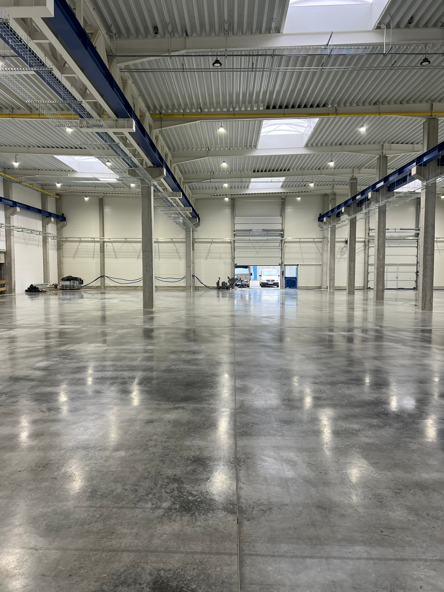 Wnętrze hali przemysłowej z betonową posadzką, widoczne słupy konstrukcyjne, instalacje pod sufitem i otwarte wrota z pojazdem w tle.