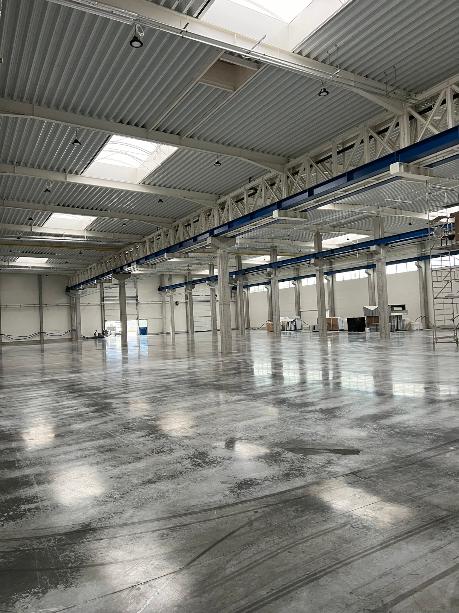 Rozległa hala przemysłowa z gładką, szarą posadzką, wsparta na rzędzie betonowych kolumn, oświetlona naturalnym światłem wpadającym przez świetliki dachowe.