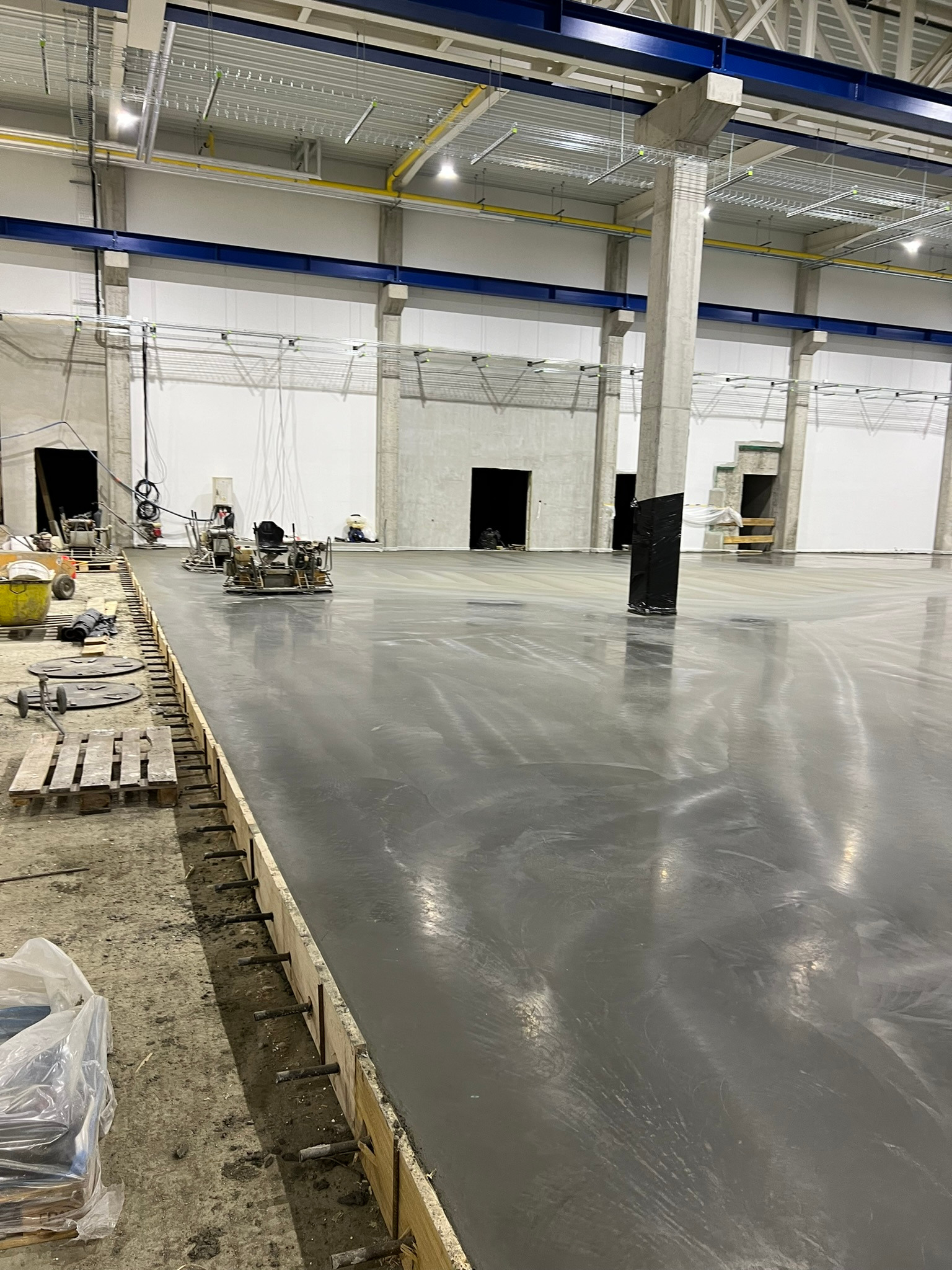Wnętrze hali przemysłowej w trakcie wykonywania posadzki, widoczne maszyny do zacierania betonu, szalunki z wystającymi prętami oraz gładka, szara powierzchnia wylewki.
