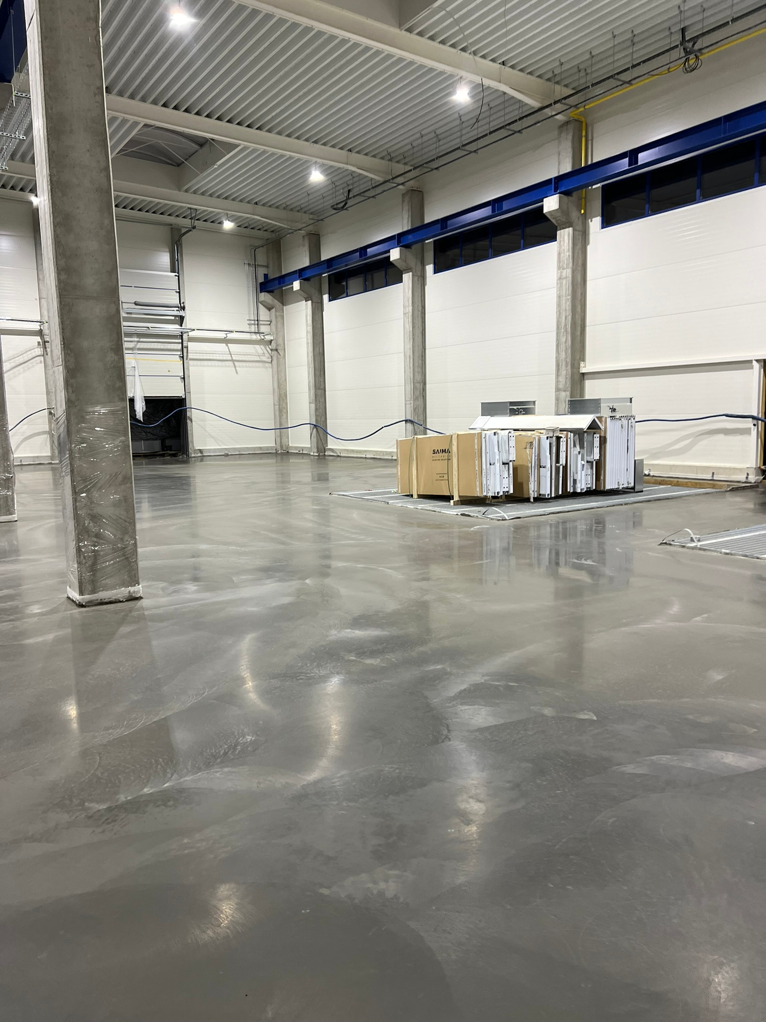 Błyszcząca, szara posadzka przemysłowa w hali produkcyjnej, z widocznymi kolumnami zabezpieczonymi folią i paletami z oknami lub drzwiami w tle.