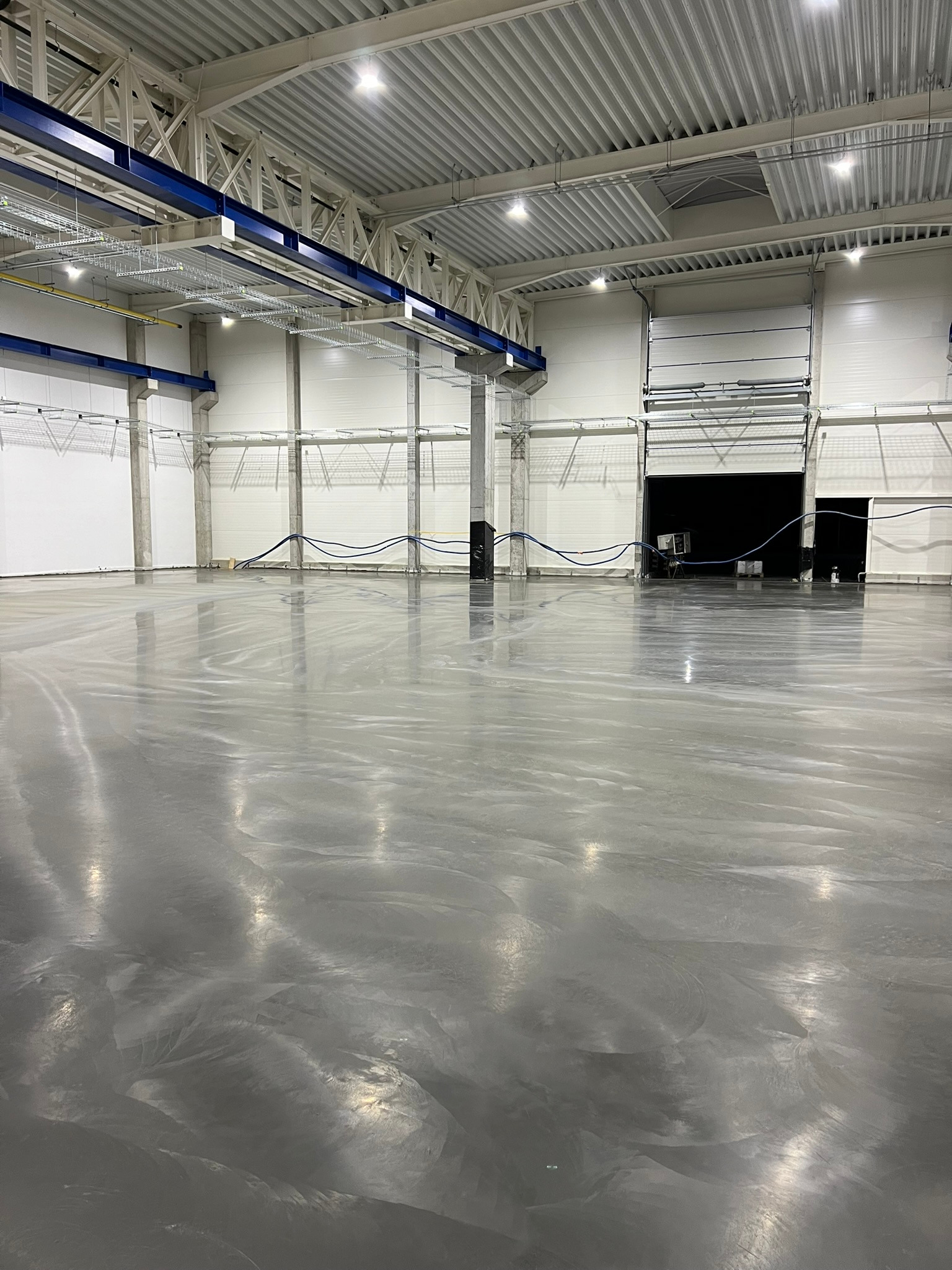 Błyszcząca, szara posadzka w hali przemysłowej, widoczne betonowe kolumny i instalacje pod sufitem, brama wjazdowa w tle.