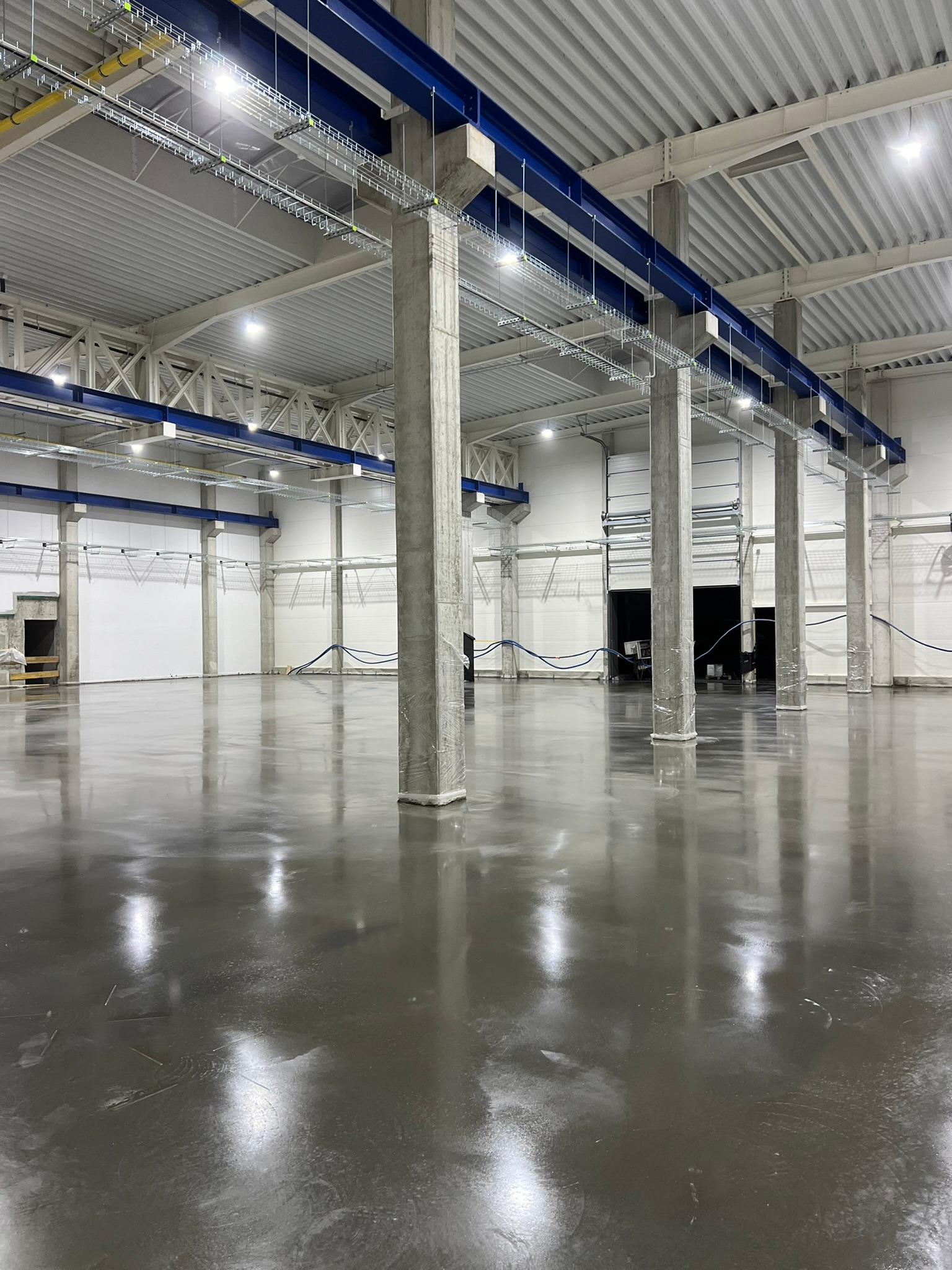 Szara, błyszcząca posadzka w hali przemysłowej, z betonowymi kolumnami i sufitową konstrukcją podtrzymującą instalacje.