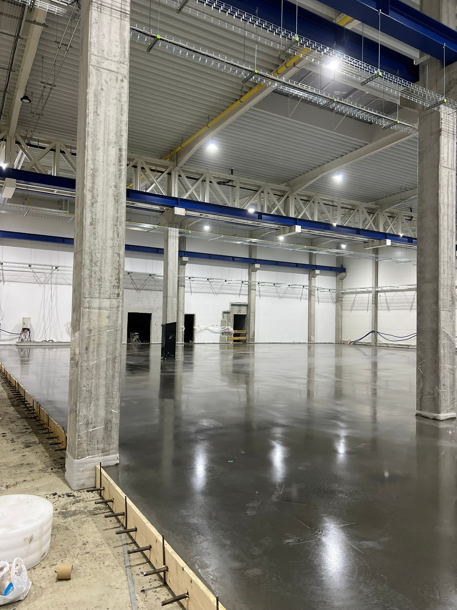 Błyszcząca, szara posadzka w hali przemysłowej, z widocznymi betonowymi filarami i elementami szalunku przy krawędzi. Konstrukcja dachu z widocznymi instalacjami.