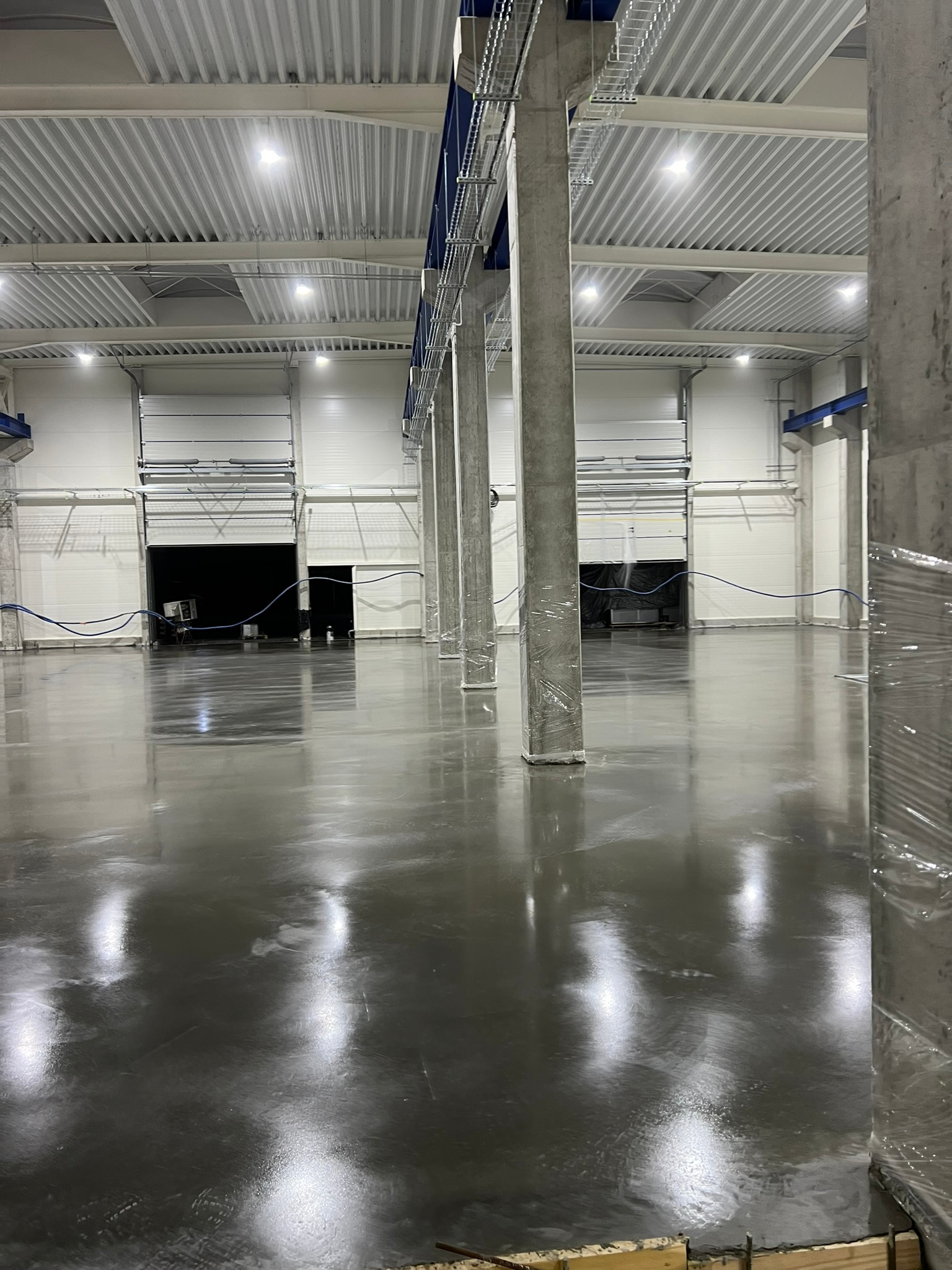 Błyszcząca, szara posadzka przemysłowa w nowej hali z widocznymi słupami owiniętymi folią ochronną i otwartymi bramami w tle.