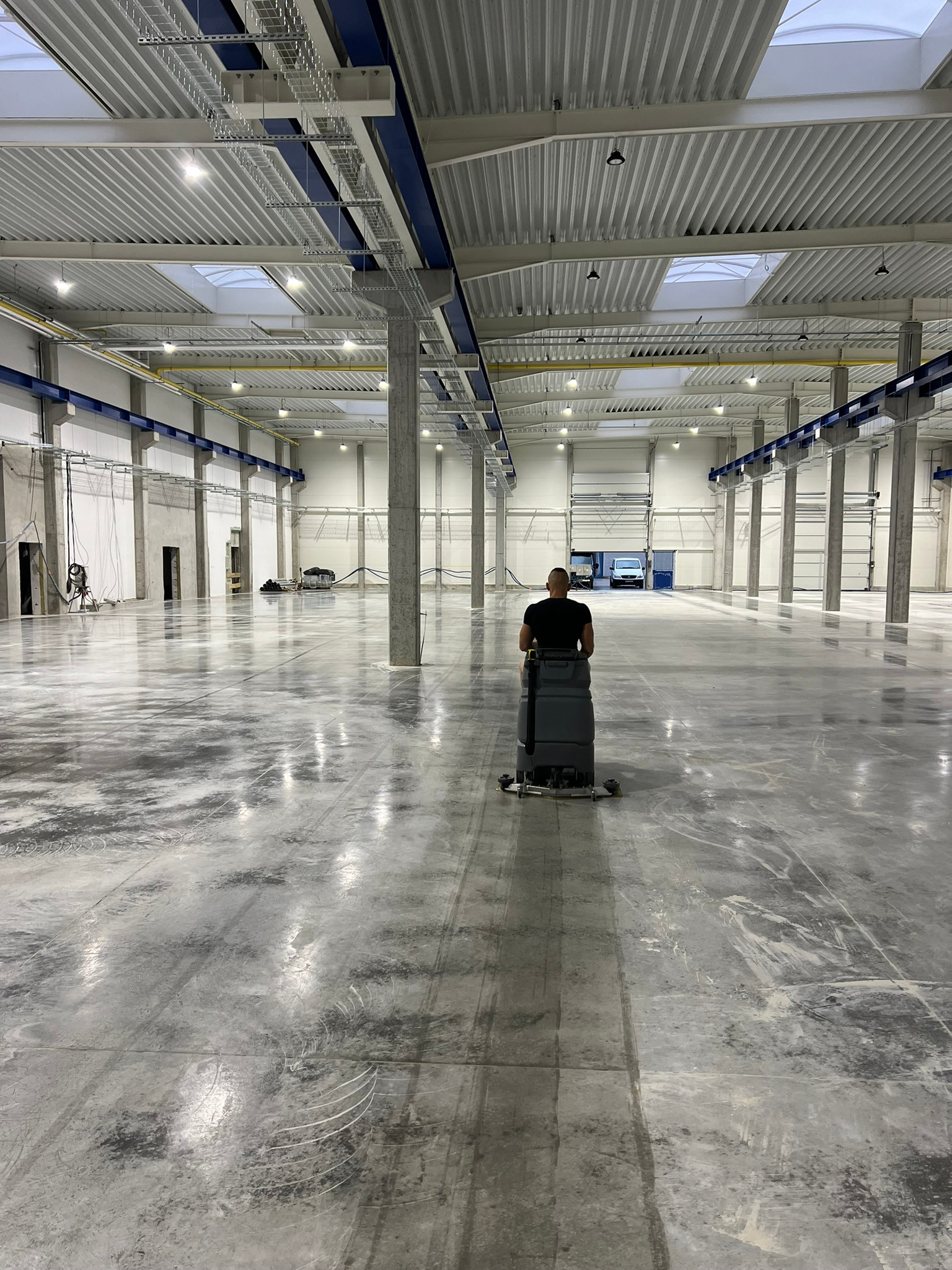 Pracownik obsługuje maszynę czyszczącą posadzkę w przestronnej hali przemysłowej z betonowymi filarami i oświetleniem sufitowym, widoczne ślady czyszczenia na szarej powierzchni.