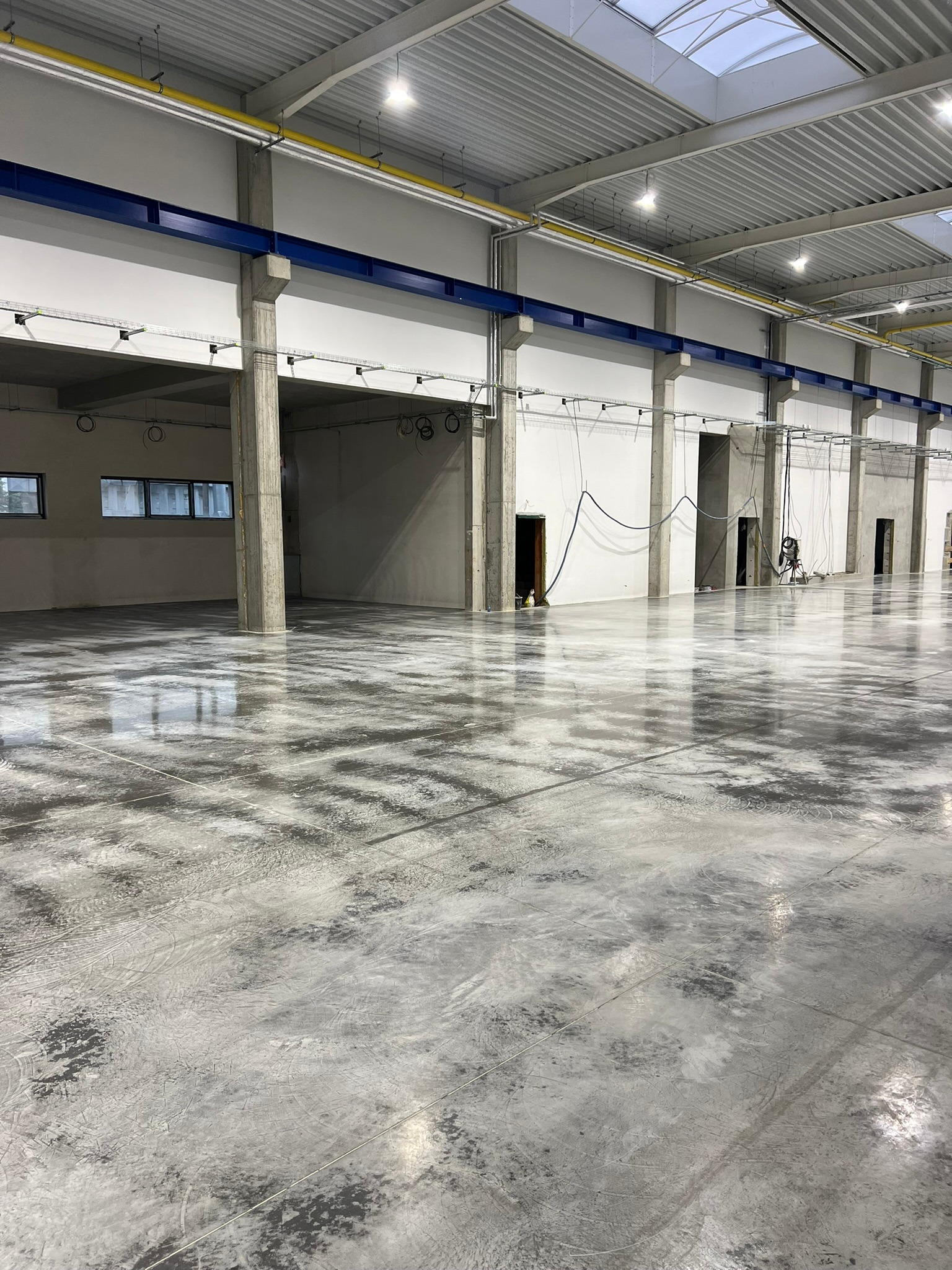 Wnętrze hali przemysłowej z betonową posadzką o szarej barwie i widocznym wzorze, oświetlonej światłem dziennym z okien dachowych i lampami sufitowymi.
