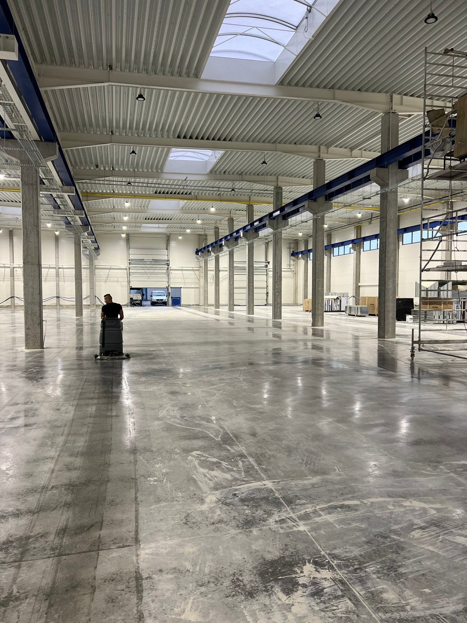 Duża hala przemysłowa z betonową posadzką w trakcie mycia maszyną czyszczącą, widoczne ślady po czyszczeniu, wysokie sufity z oknami dachowymi i betonowe kolumny.