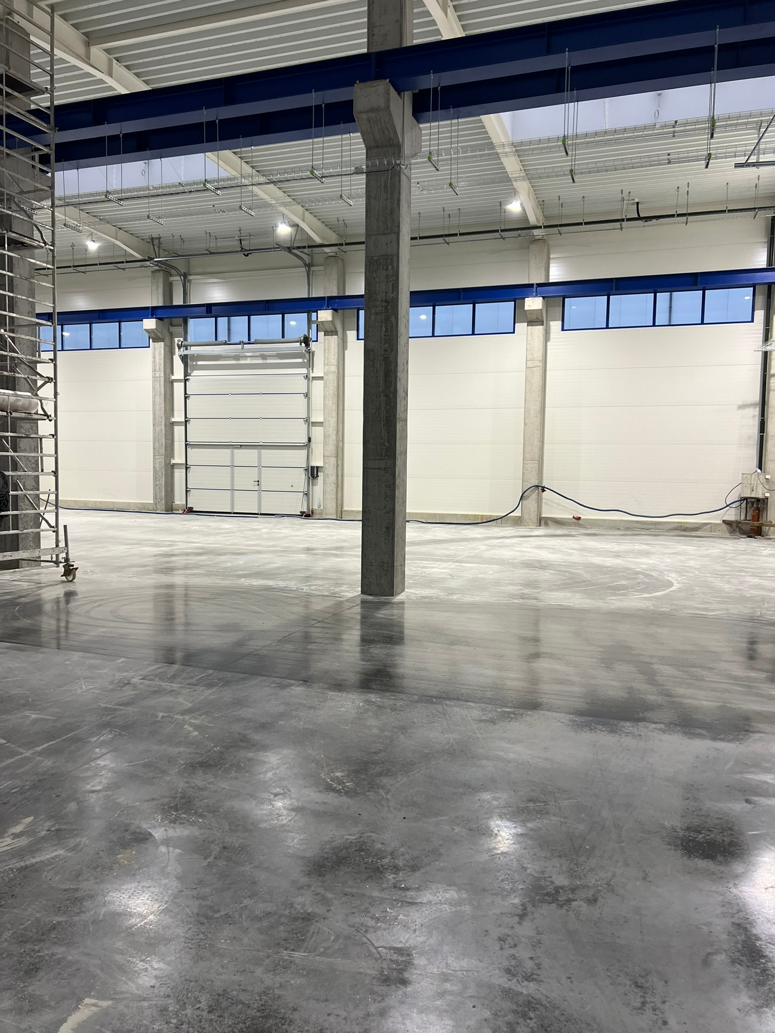 Wnętrze hali przemysłowej z betonową posadzką, widoczne słupy konstrukcyjne, brama wjazdowa oraz metalowe rusztowanie z boku.
