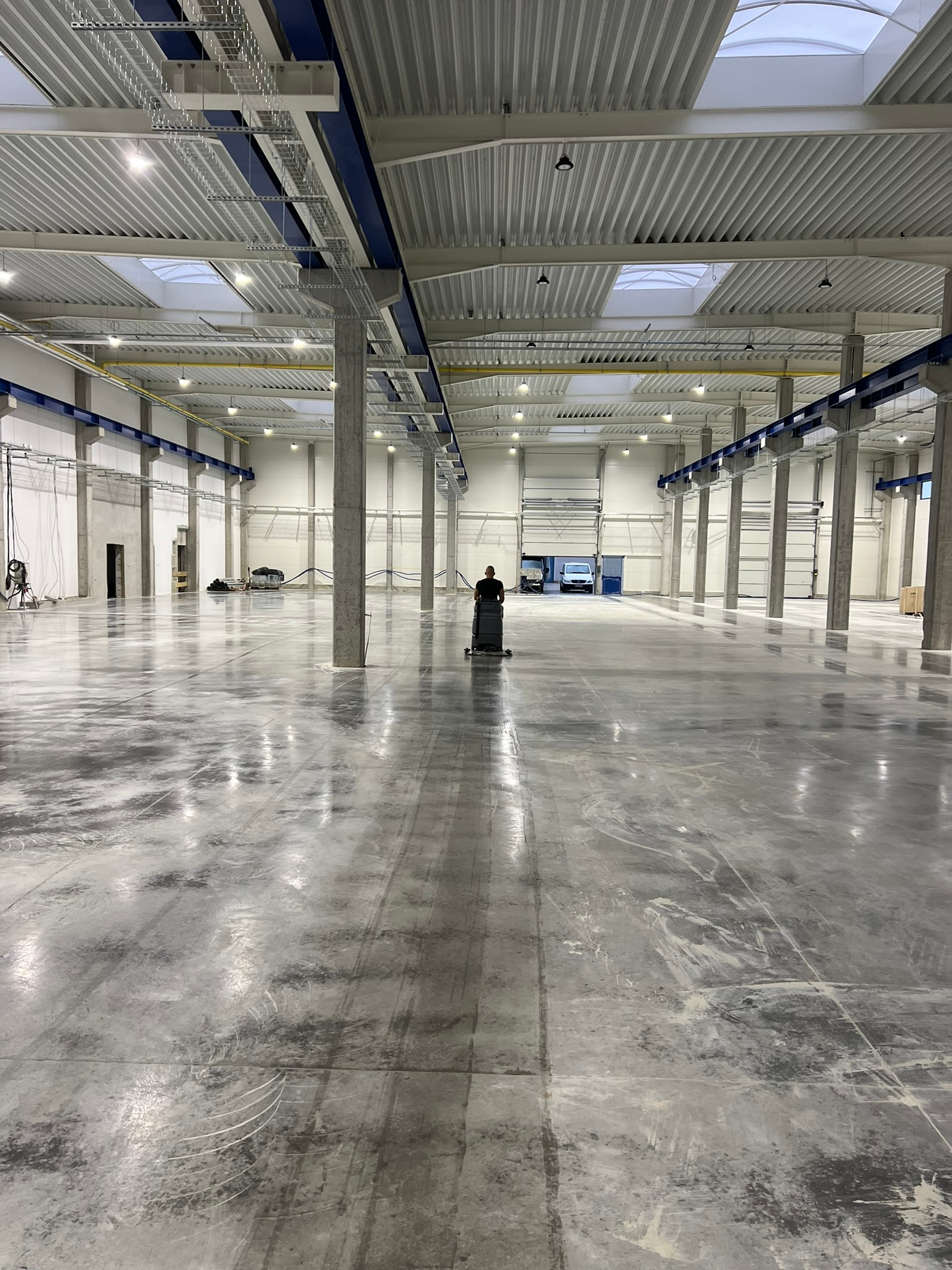 Pracownik obsługuje maszynę czyszczącą na dużej, szarej powierzchni w hali przemysłowej z betonowymi kolumnami i świetlikami w suficie.