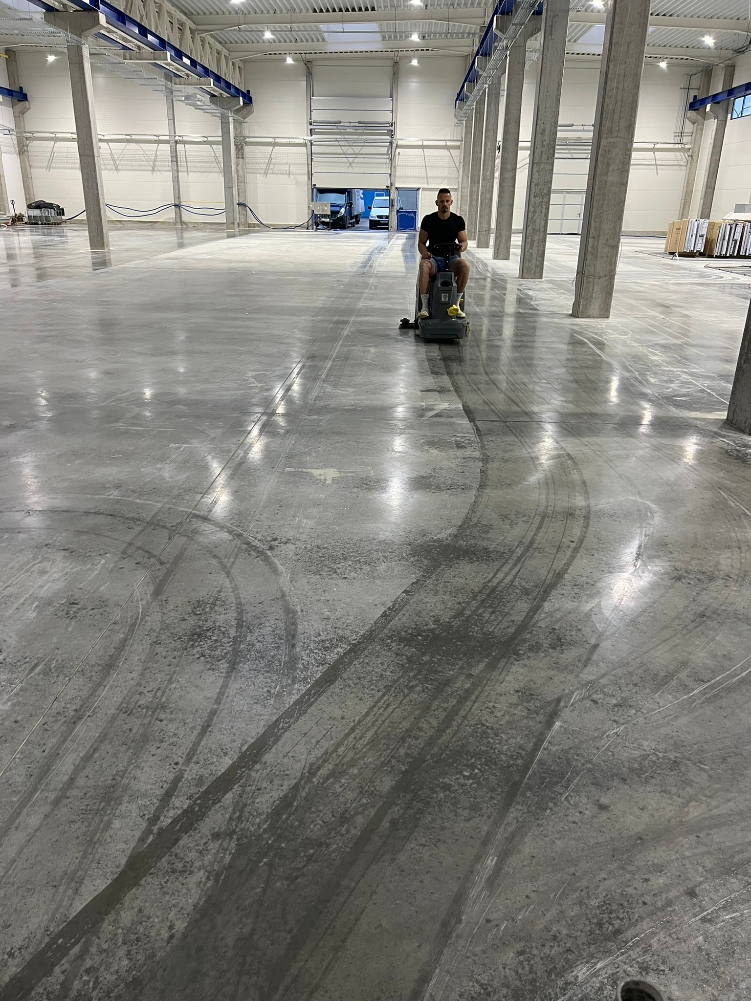 Pracownik na maszynie czyszczącej poleruje betonową posadzkę w hali przemysłowej, widoczne ślady po czyszczeniu.