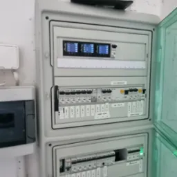 Otwarta skrzynka rozdzielcza z zainstalowanymi bezpiecznikami i modułami sterującymi, z routerem Wi-Fi umieszczonym na górze, na tle białej ściany.